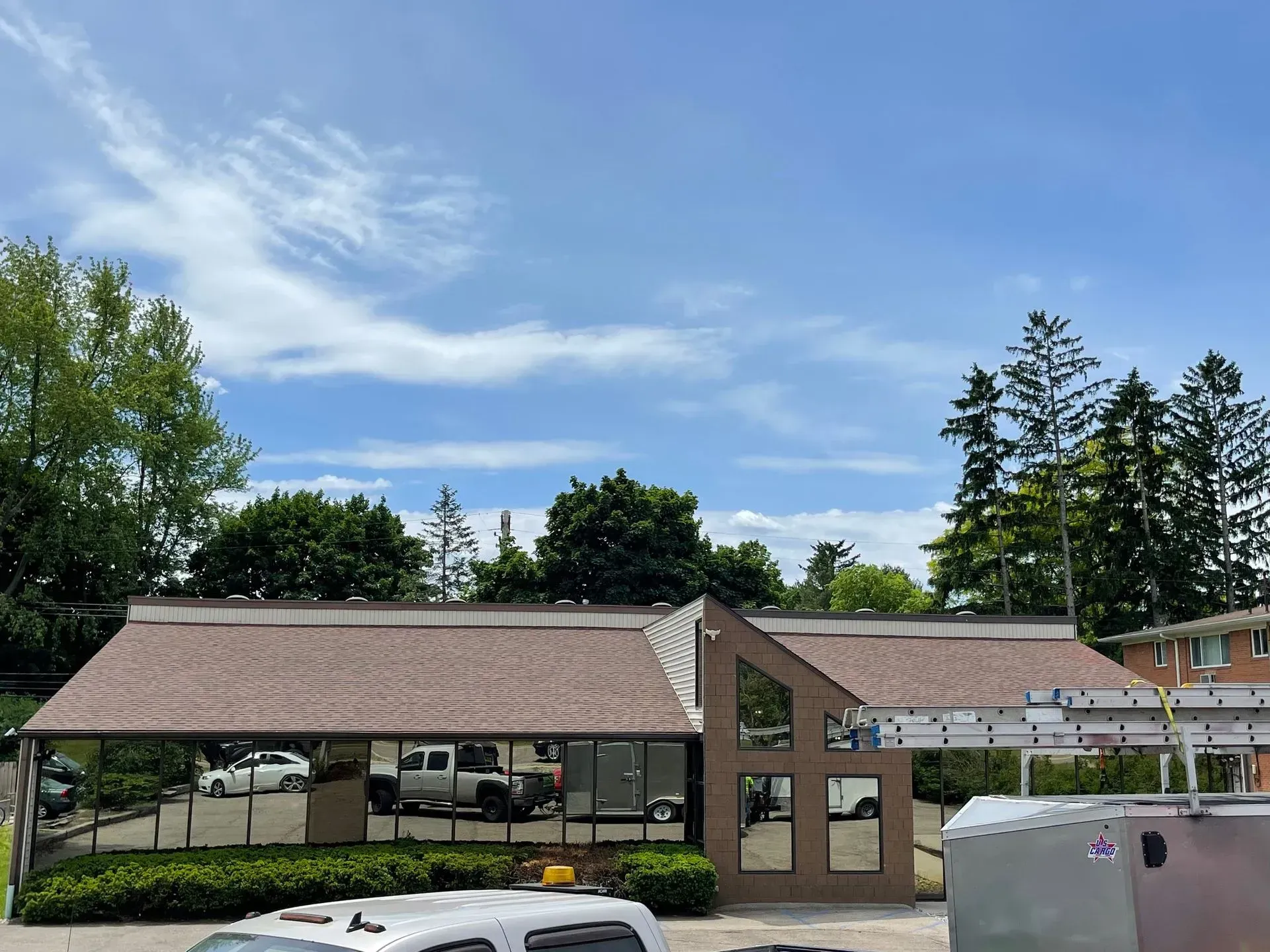 Single-story building with tan roofs and large windows under a blue sky, surrounded by trees and a parking lot.