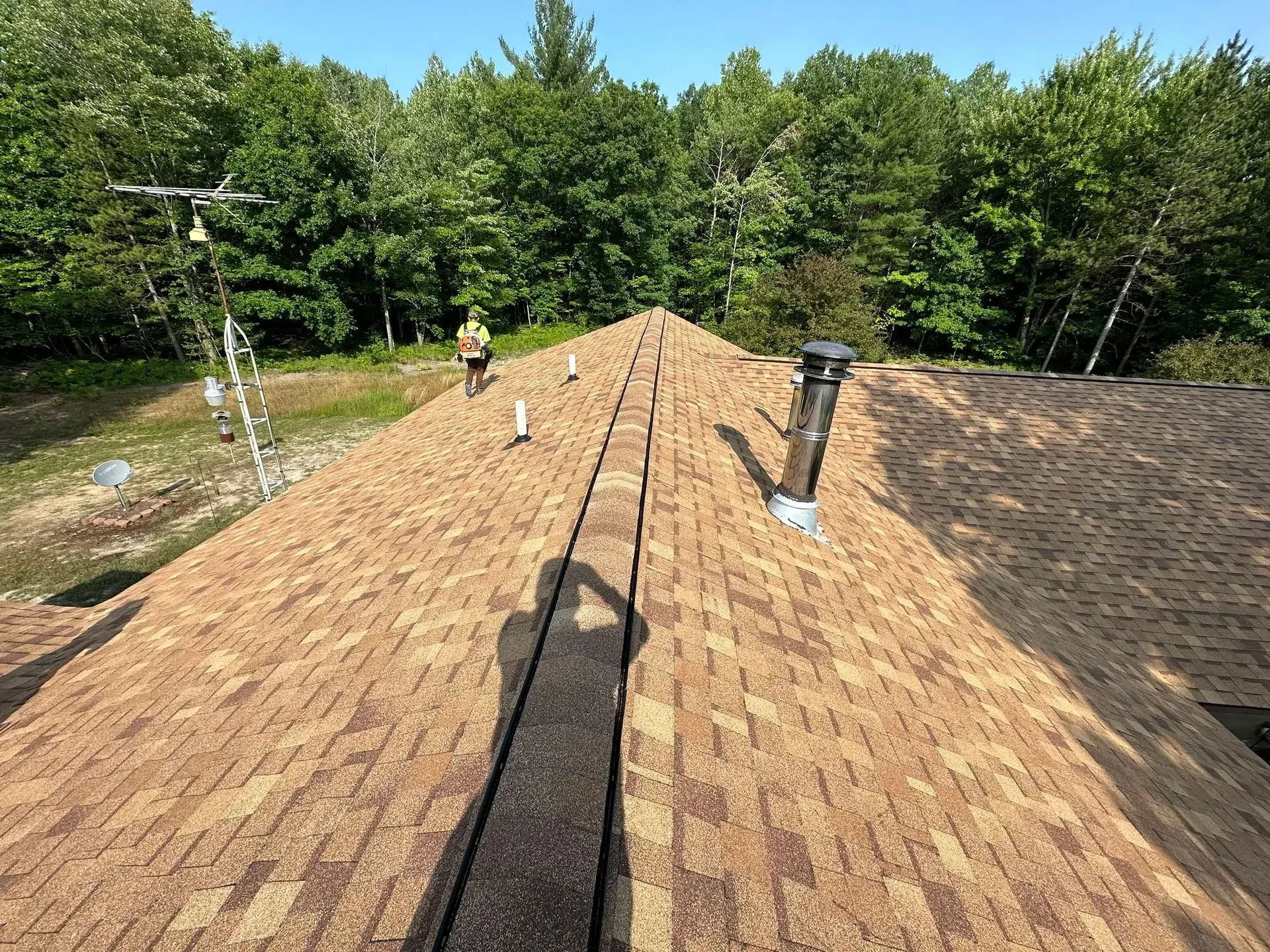 Shingled roof with vents and a worker near the ridge, surrounded by trees under a clear sky