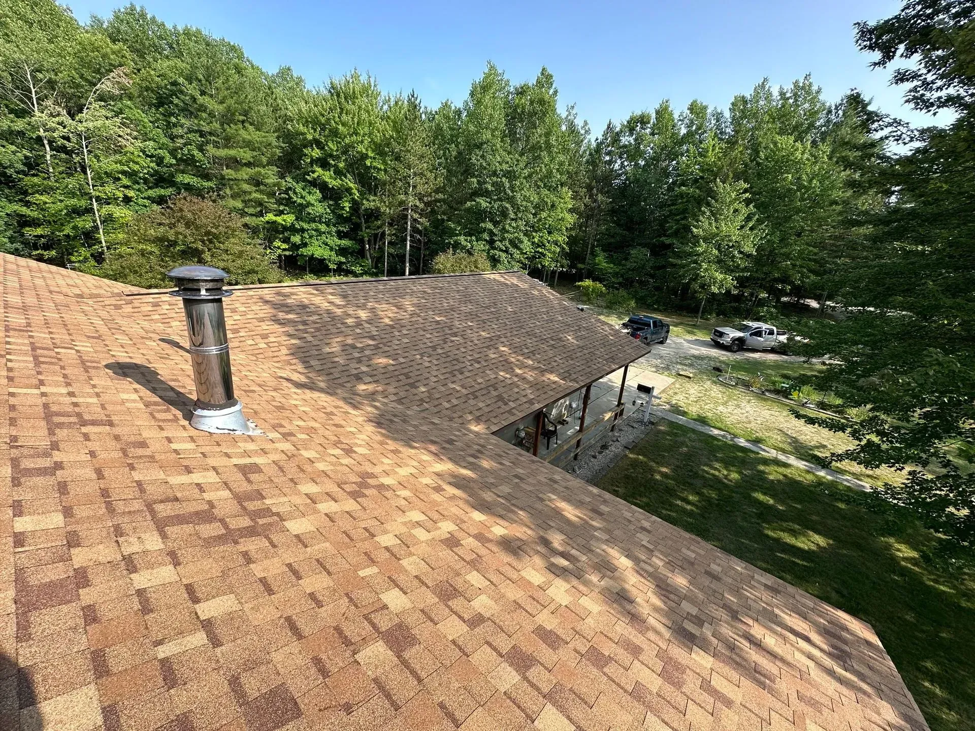 Shingled roof with vent under a clear sky, surrounded by green trees and a grassy yard