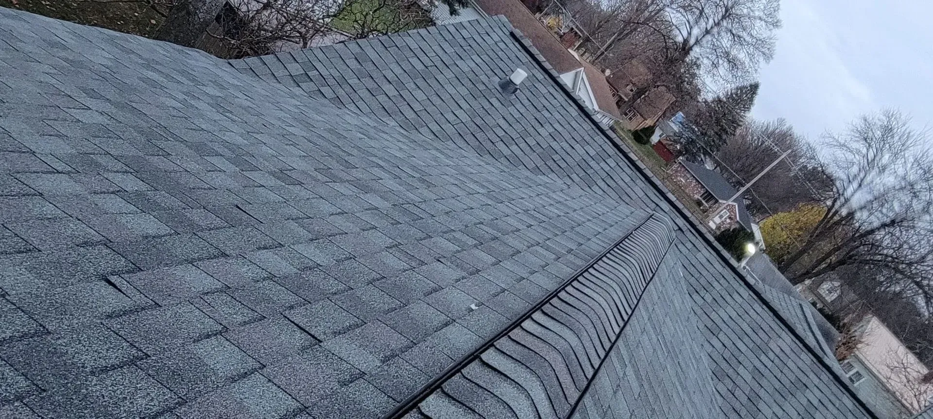 Rooftop covered in gray shingles with a metal ridge vent, viewed diagonally under an overcast sky