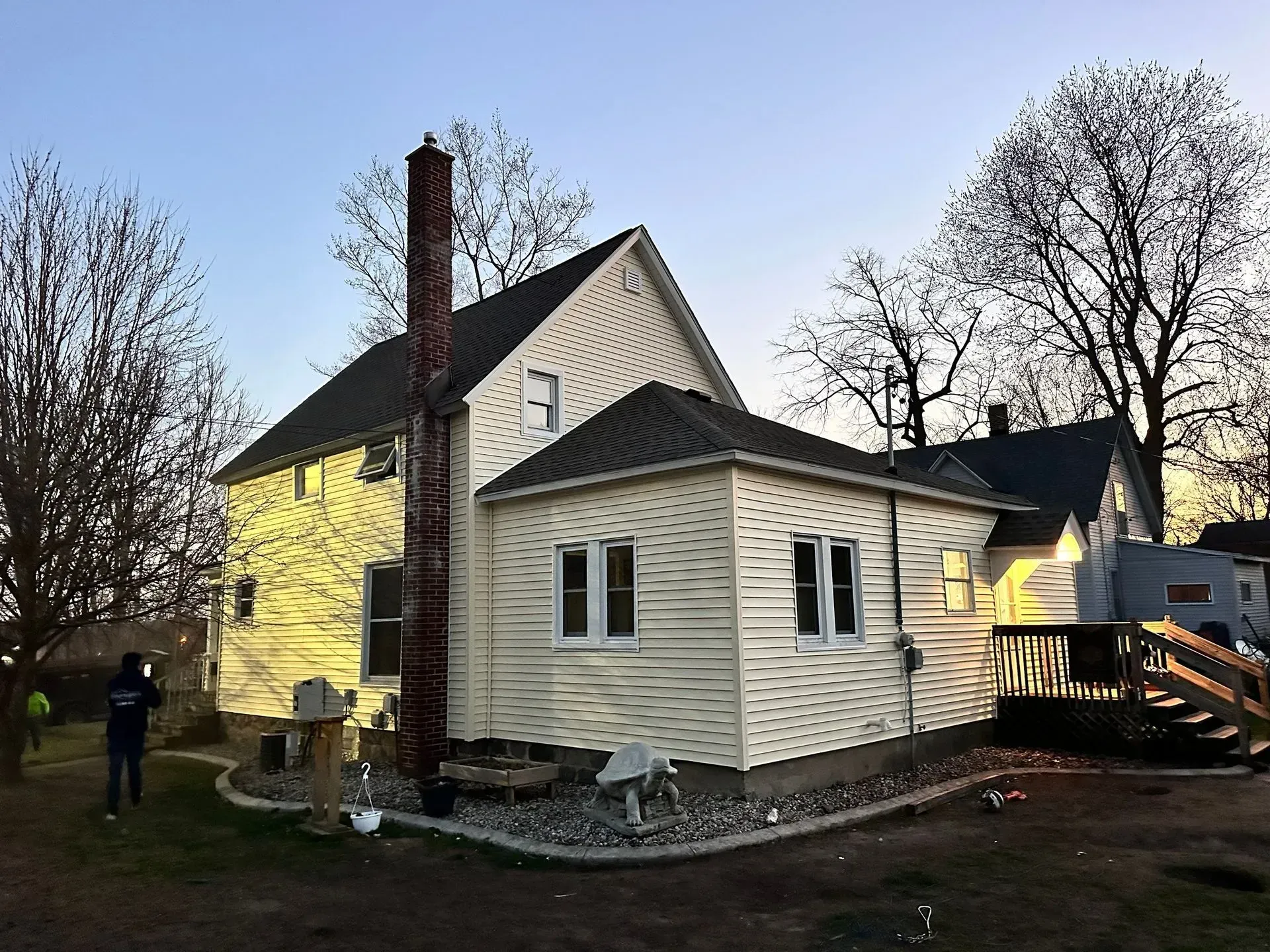 House with white siding and brick chimney at dusk, with bare trees and a person near the yard