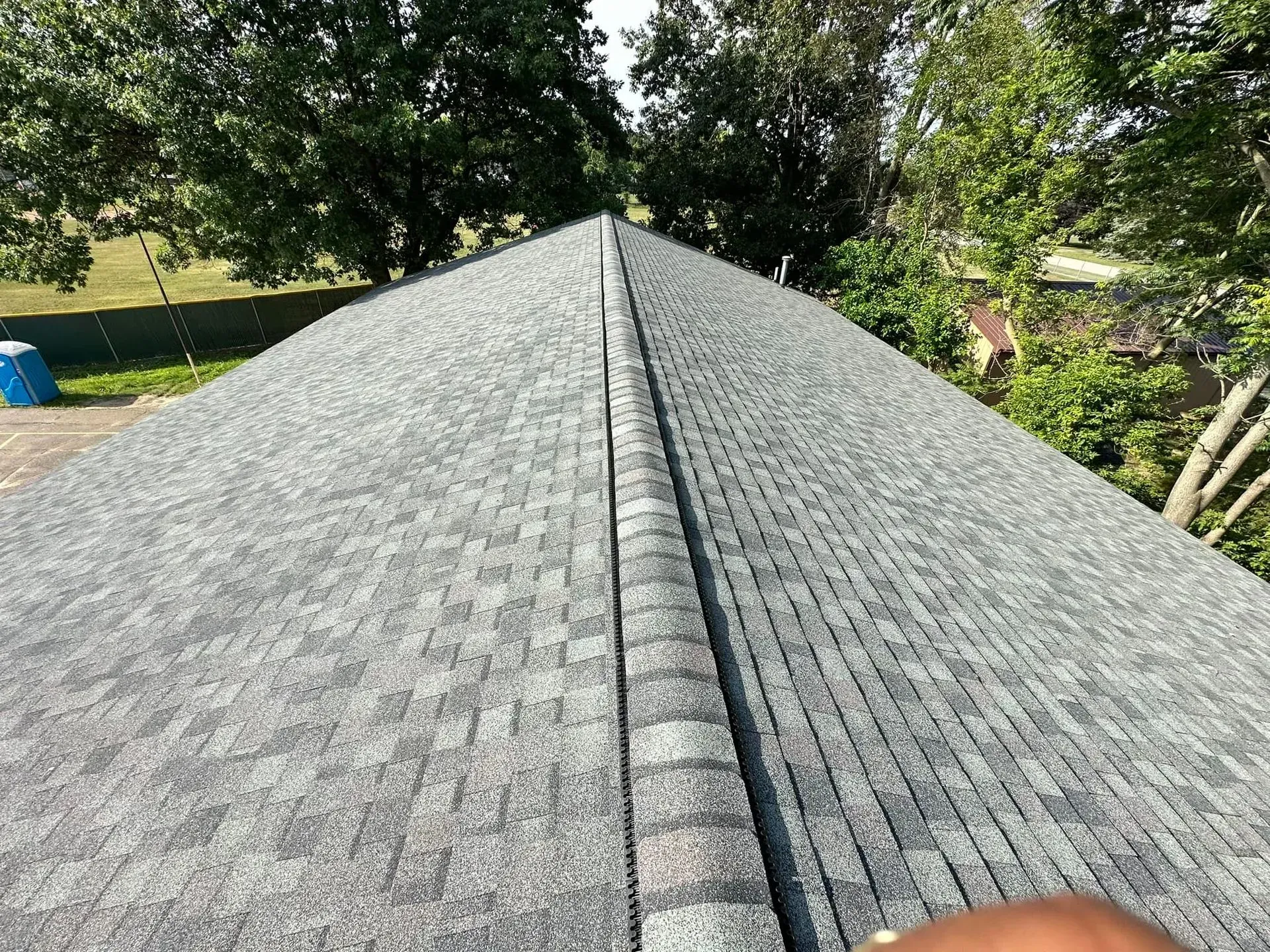 Gray shingled roof with a ridge vent, viewed from the peak over a green yard and trees