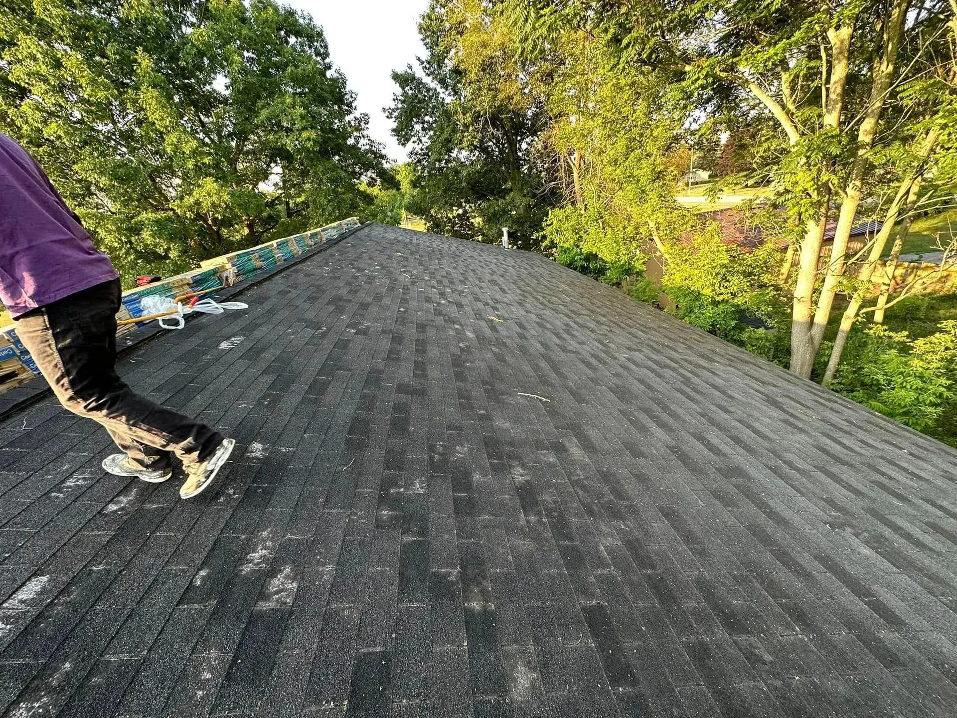 Person walking on a dark shingled roof surrounded by trees in daylight