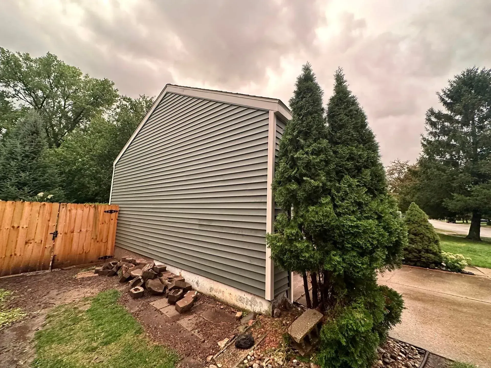 House siding buckling outward beside a wooden fence and evergreen shrubs in a yard