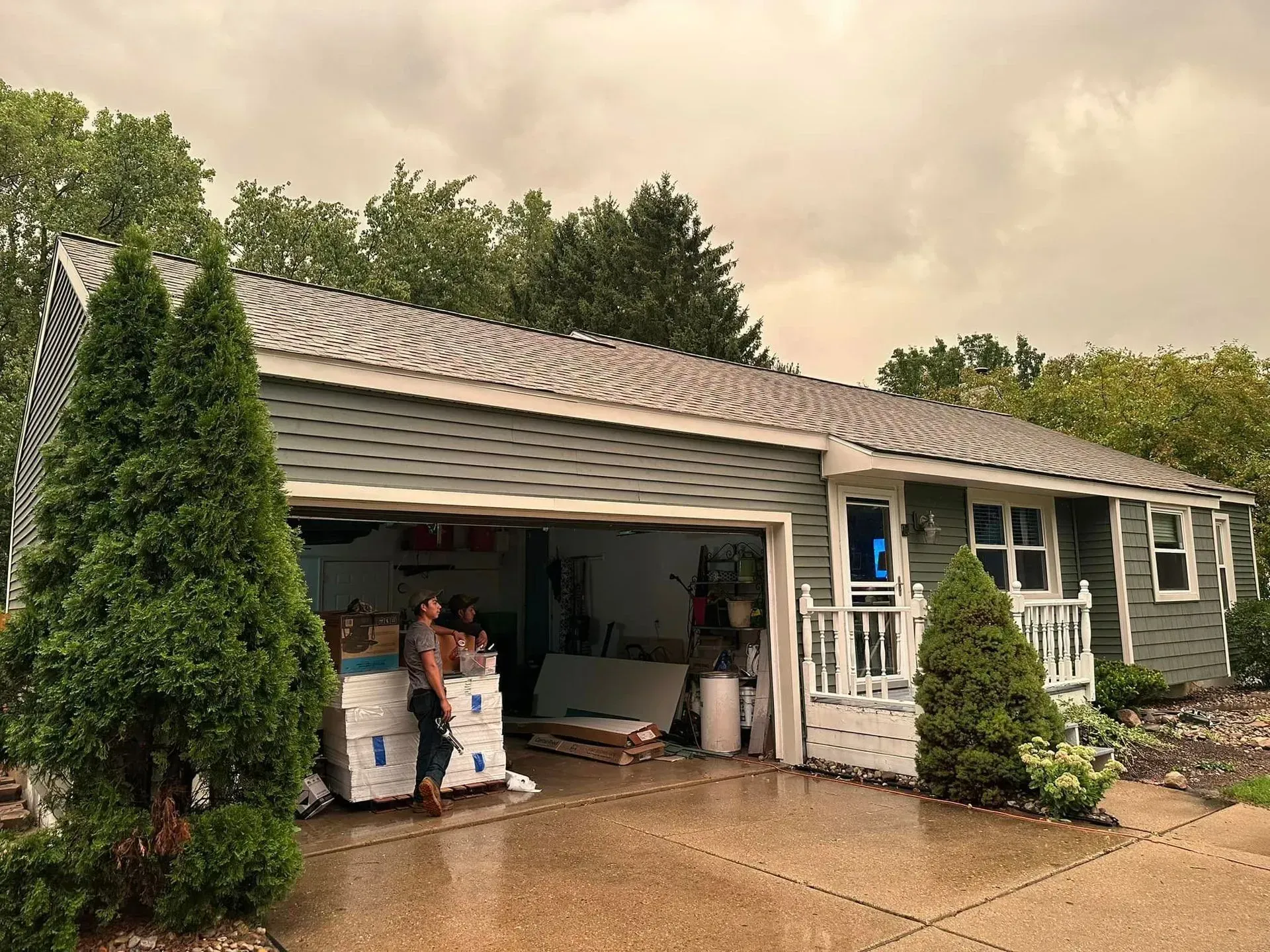 Open garage with stacked boxes and a house exterior after rain, framed by tall shrubs.