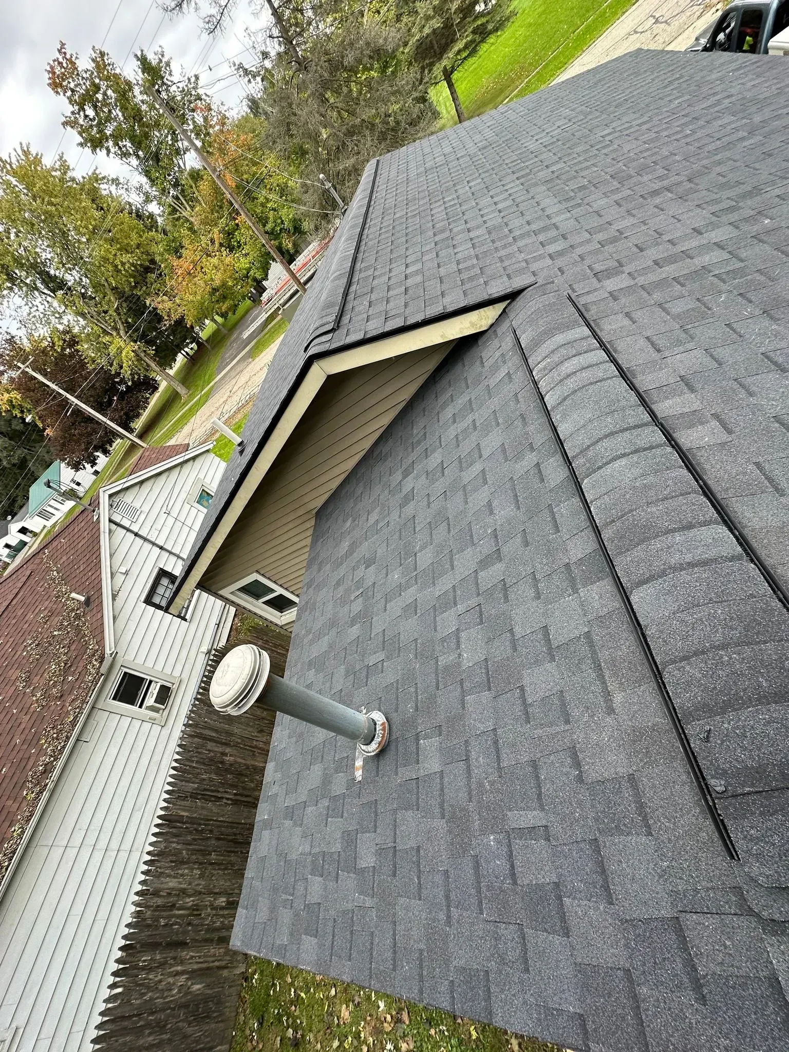 Rooftop with gray shingles, vent pipe, and brick chimney beside a white house and fenced yard.