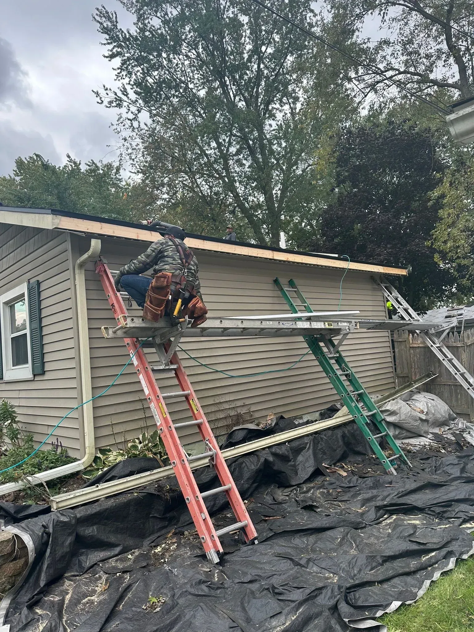 Worker on ladder repairing damaged house roof amid debris and black tarp after storm.