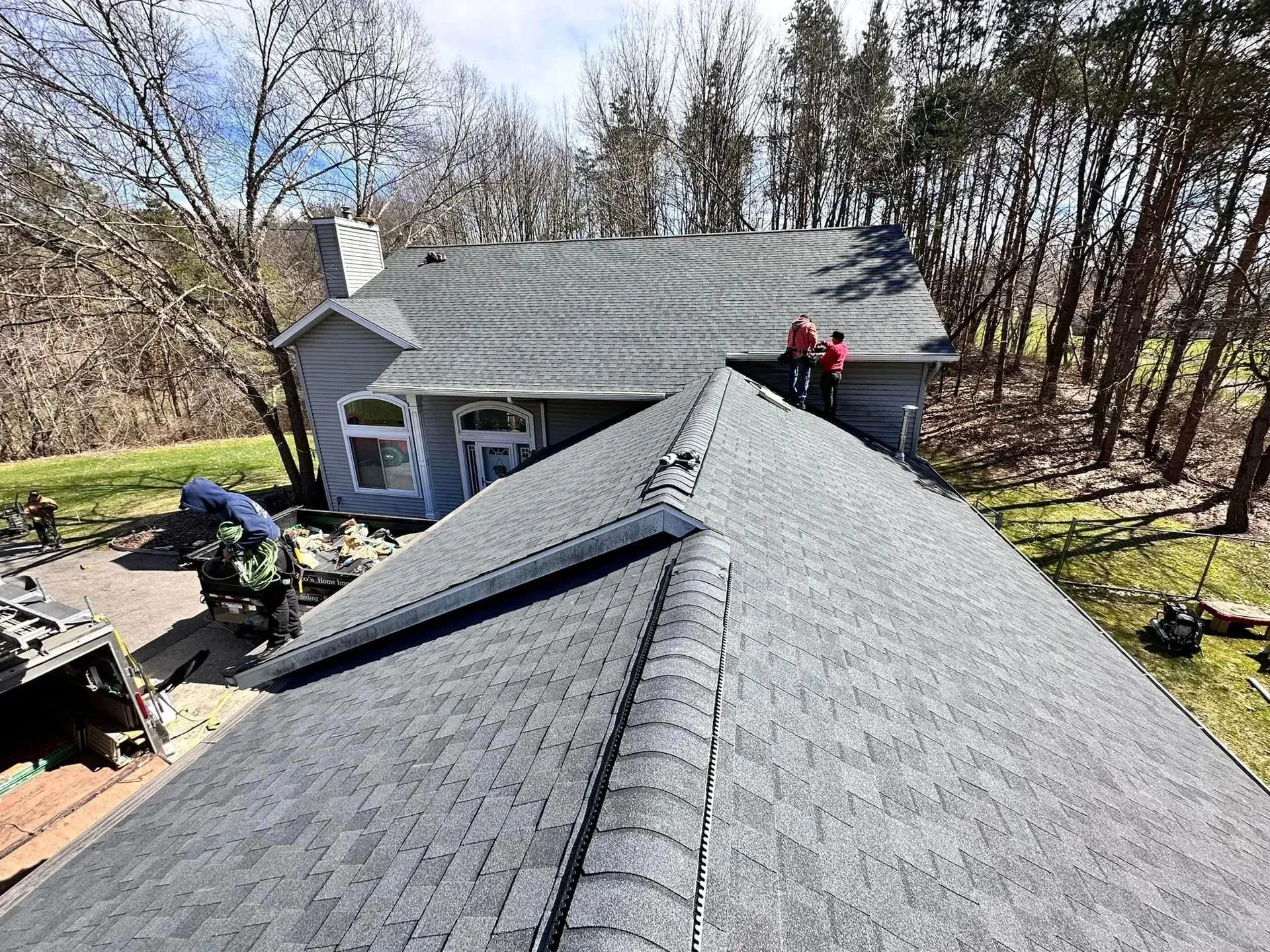 Roofers working on a gray shingled roof beside a wooded house yard