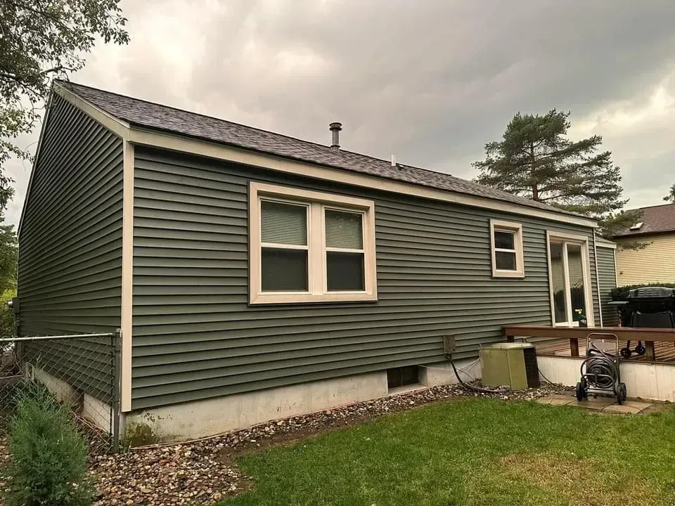 Green-sided house exterior with white-trimmed windows, grass yard, and patio area with outdoor equipment