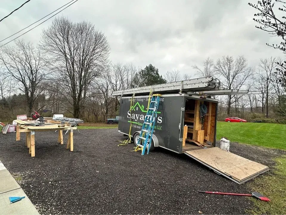 Outdoor worksite with a trailer, scattered tools, and a ladder on gravel under a cloudy sky