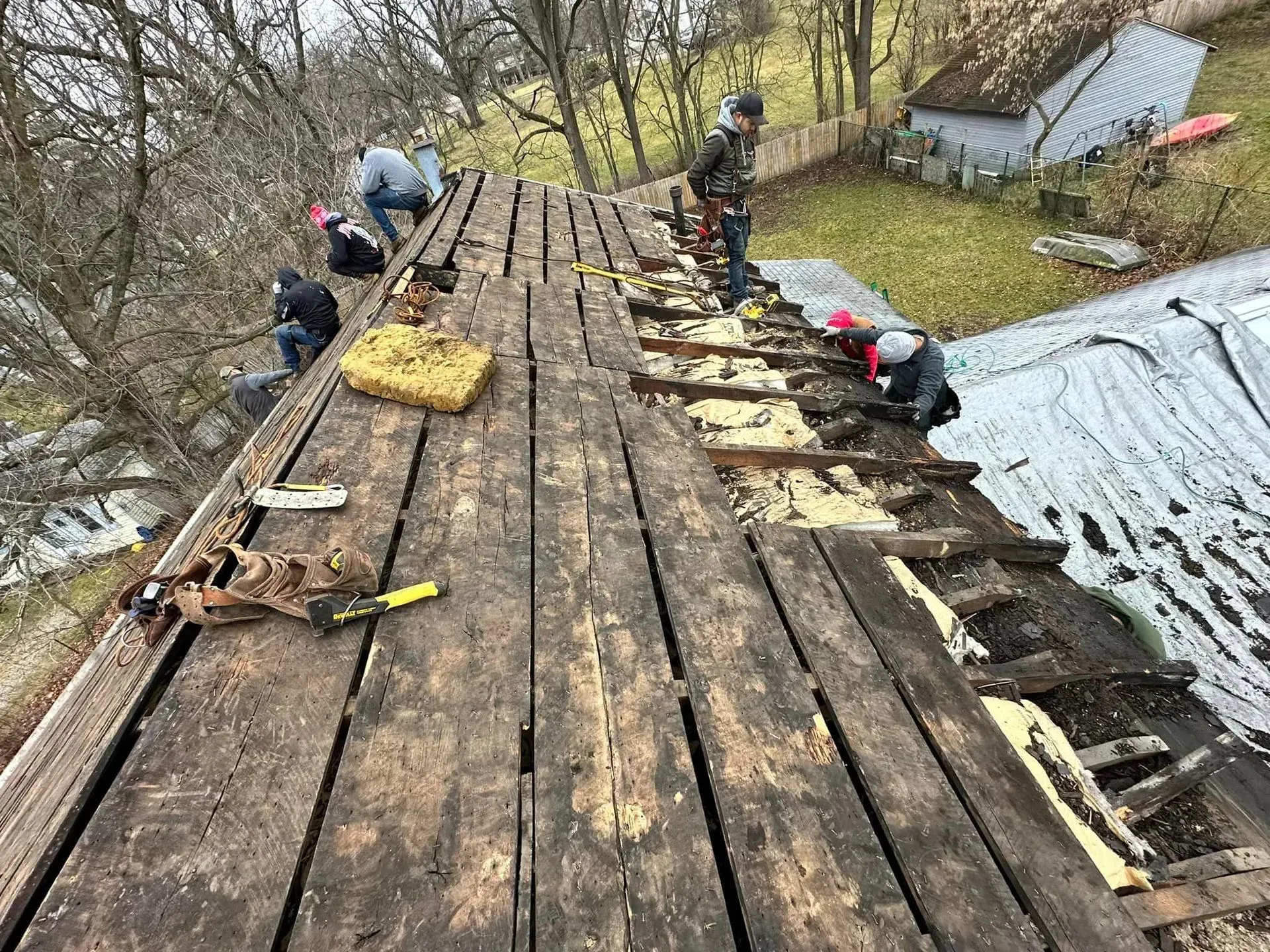 Workers repairing a worn wooden roof on a hillside cabin, with tools and tarps nearby.