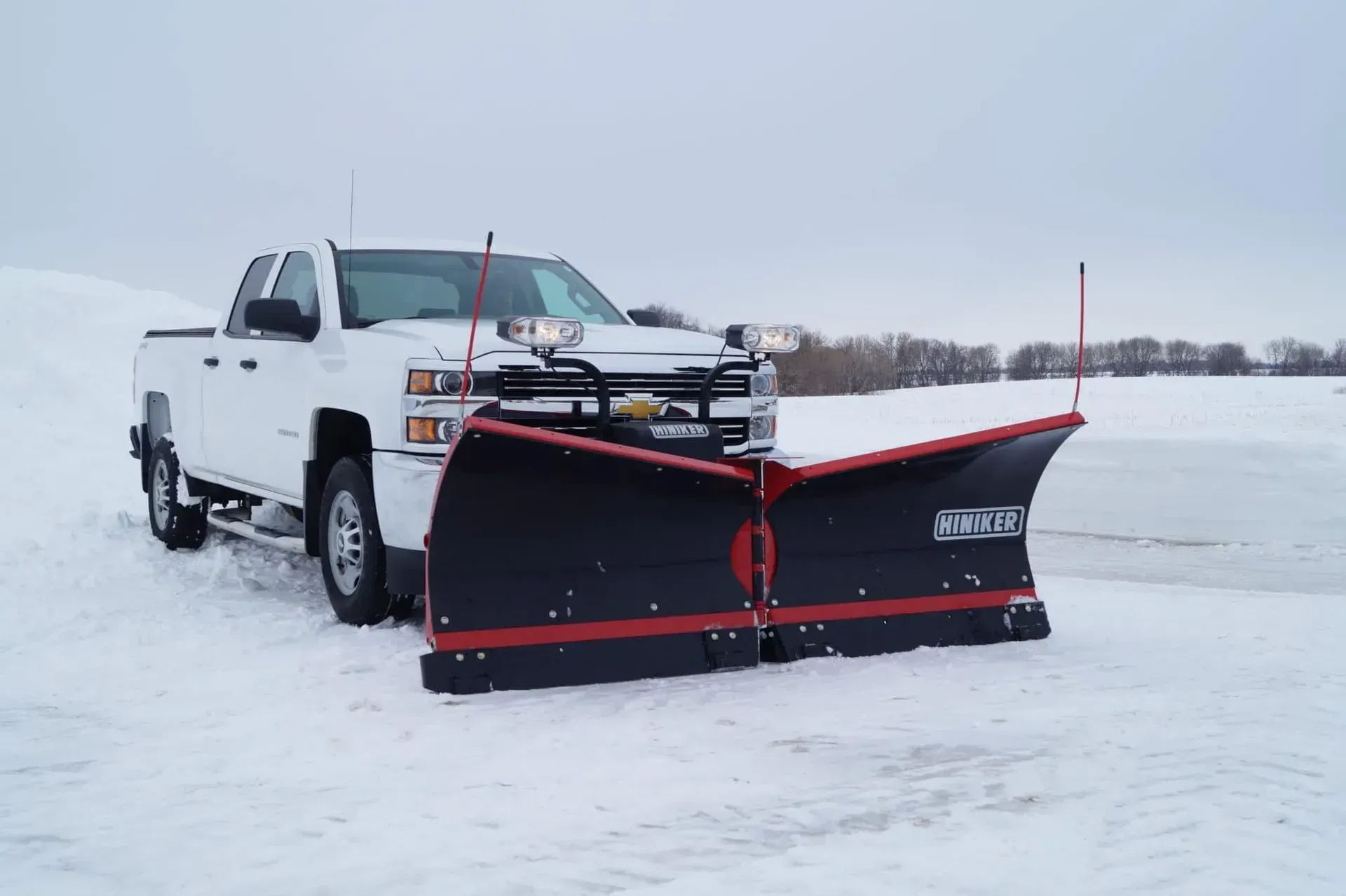 White pickup truck with a black snowplow blade on a snowy road under an overcast sky