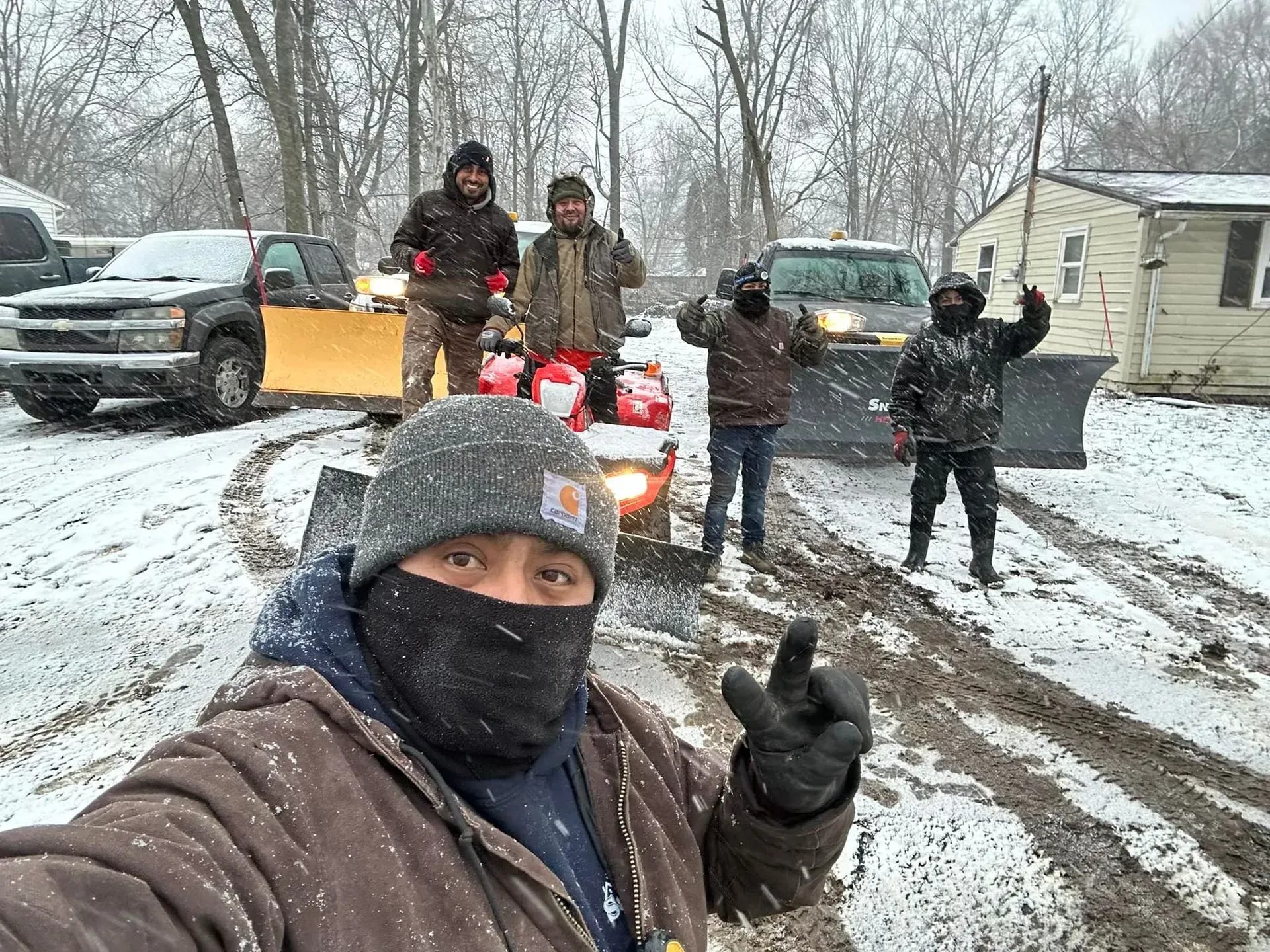 Group of people posing with shovels in a snowy driveway, one person taking a selfie in front.
