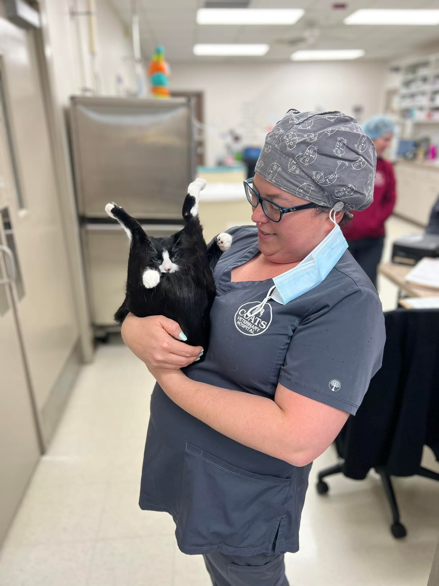 Woman in scrubs holding a black-and-white baby goat in a clinic hallway.