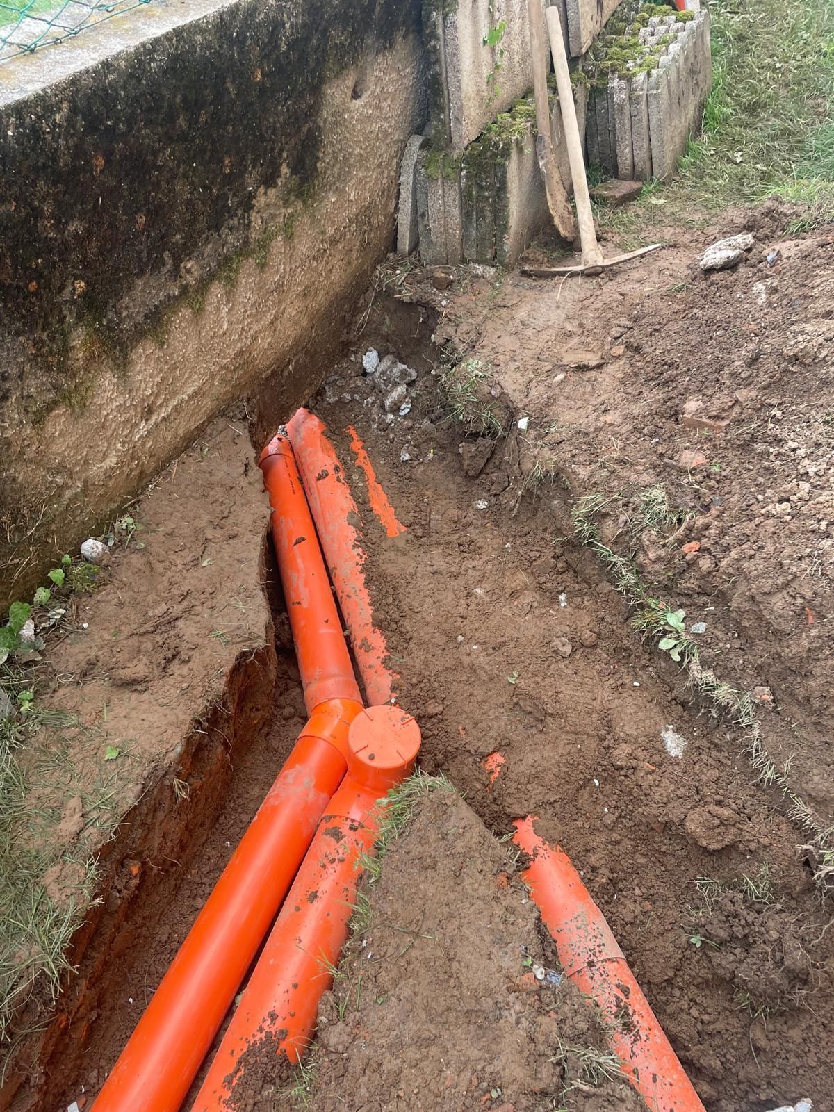 Tubature di servizio arancioni in una trincea fangosa accanto a un muro di cemento durante i lavori di costruzione.