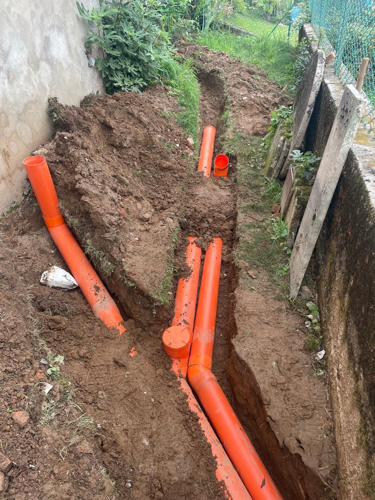 Tubi di servizio arancioni posati in una trincea fangosa accanto a un muro e una recinzione.