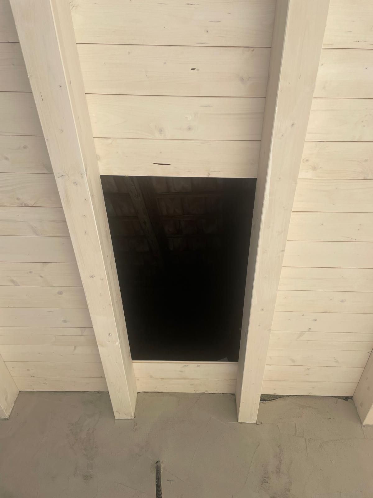 Vista di un soffitto in legno bianco con un'apertura rettangolare scura tra le travi, sopra un pavimento grigio.