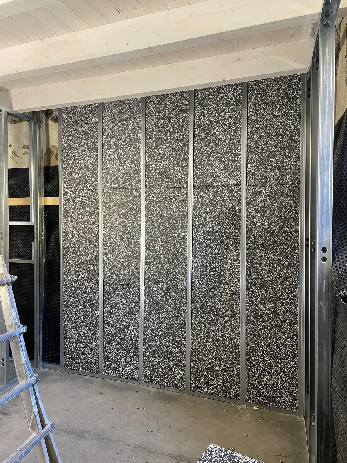 Parete interna non rifinita con pannelli isolanti grigi incorniciati da montanti metallici, accanto a una scala.