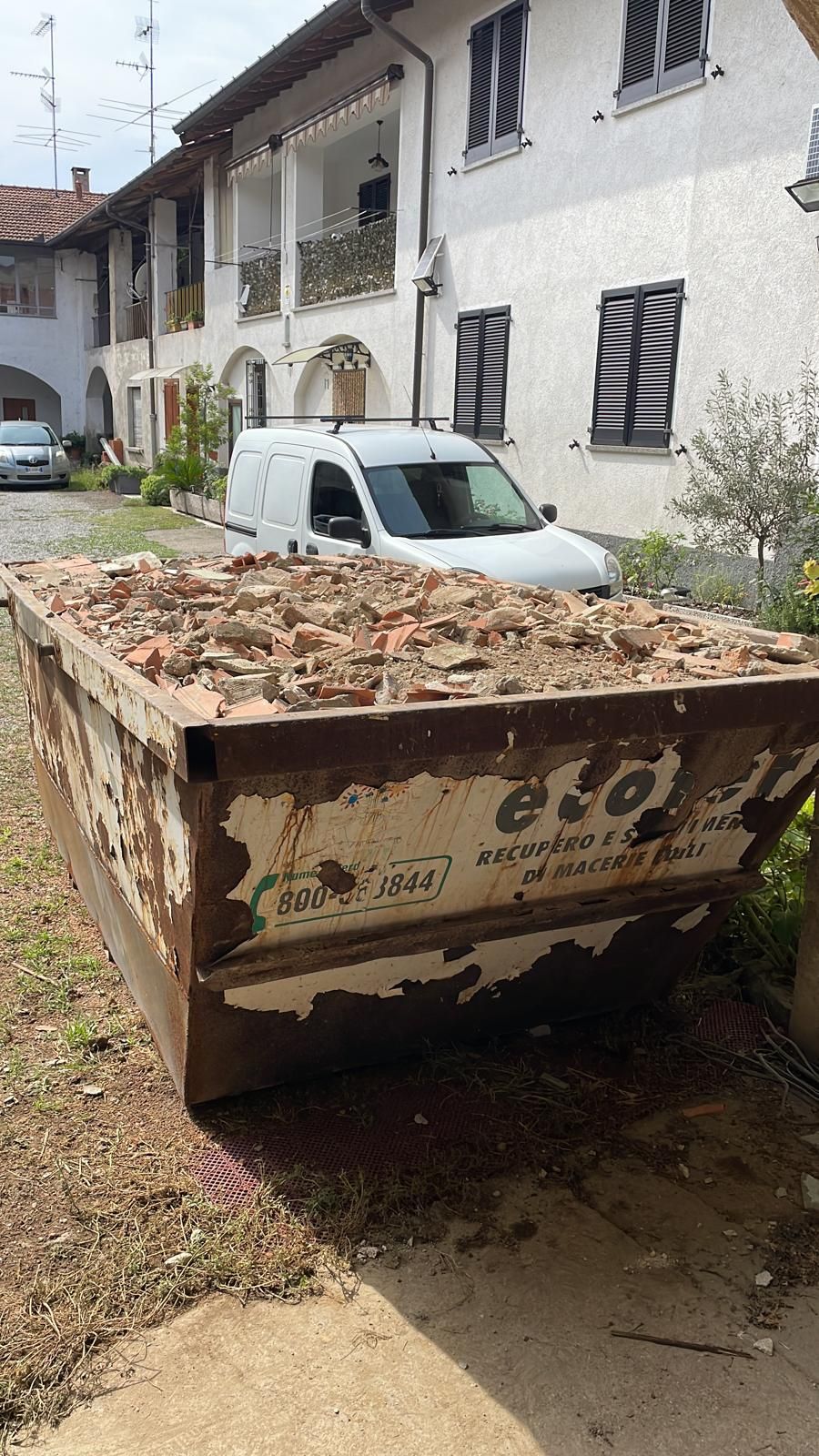 Un cassonetto pieno di detriti si trova di fronte a un edificio bianco, con un piccolo furgone parcheggiato alle sue spalle.