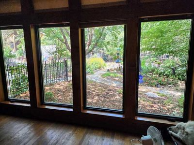 Three rectangular windows offer a view of a lush garden with a winding path. A dog and a fan are in the foreground.