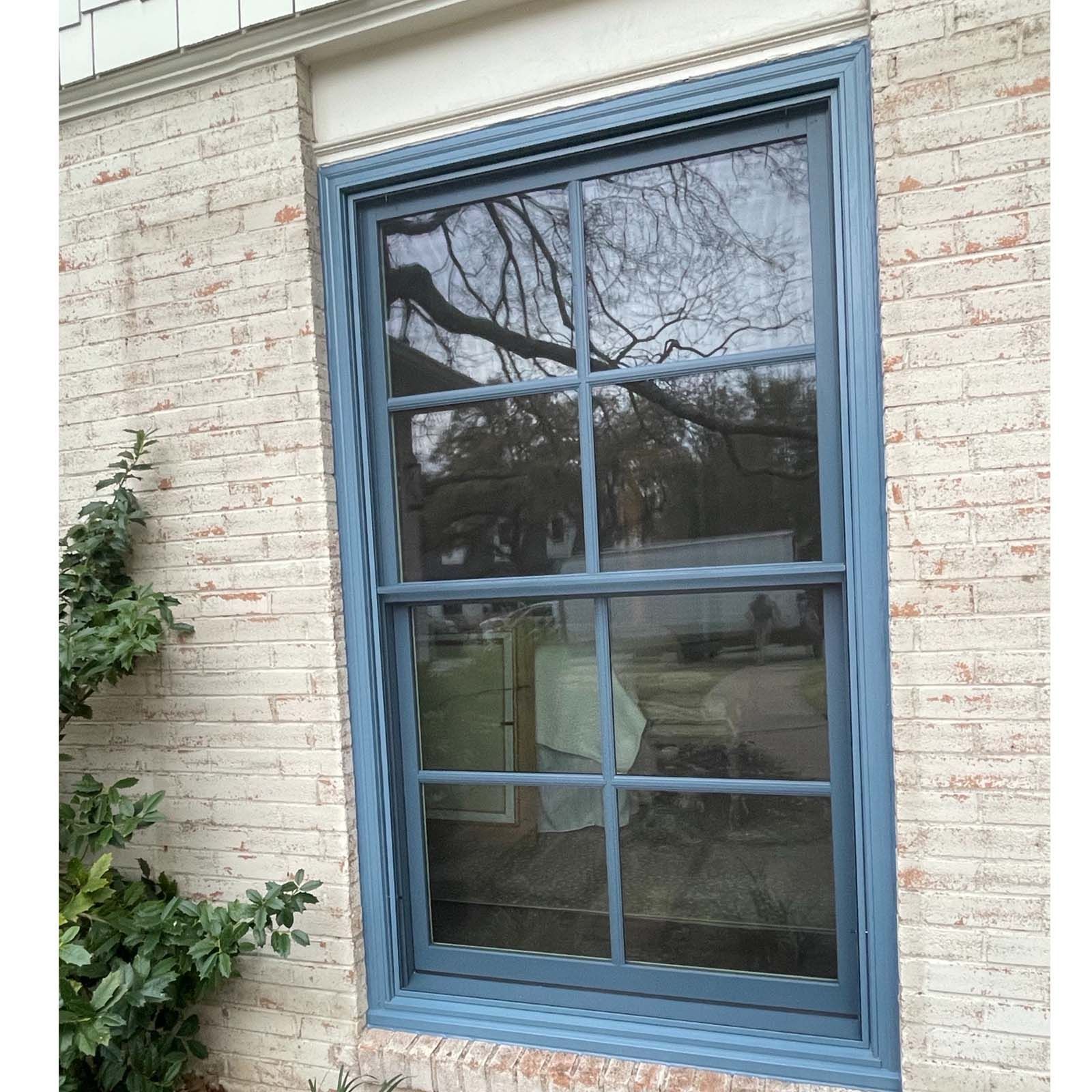 Blue-framed window with a grid pattern on a brick wall, reflecting trees.