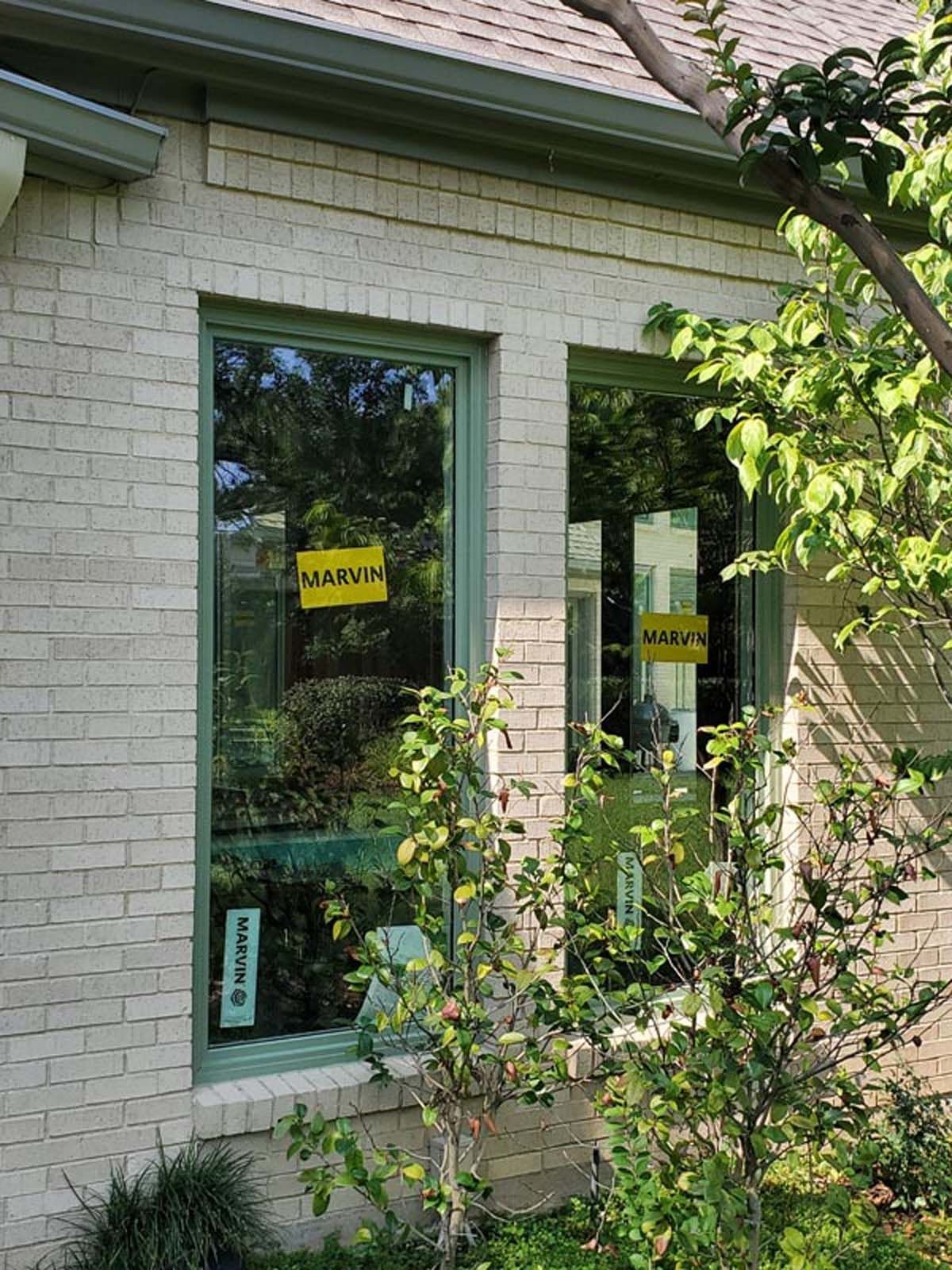 Two windows with yellow signs on a brick building with green trim and leafy greenery.