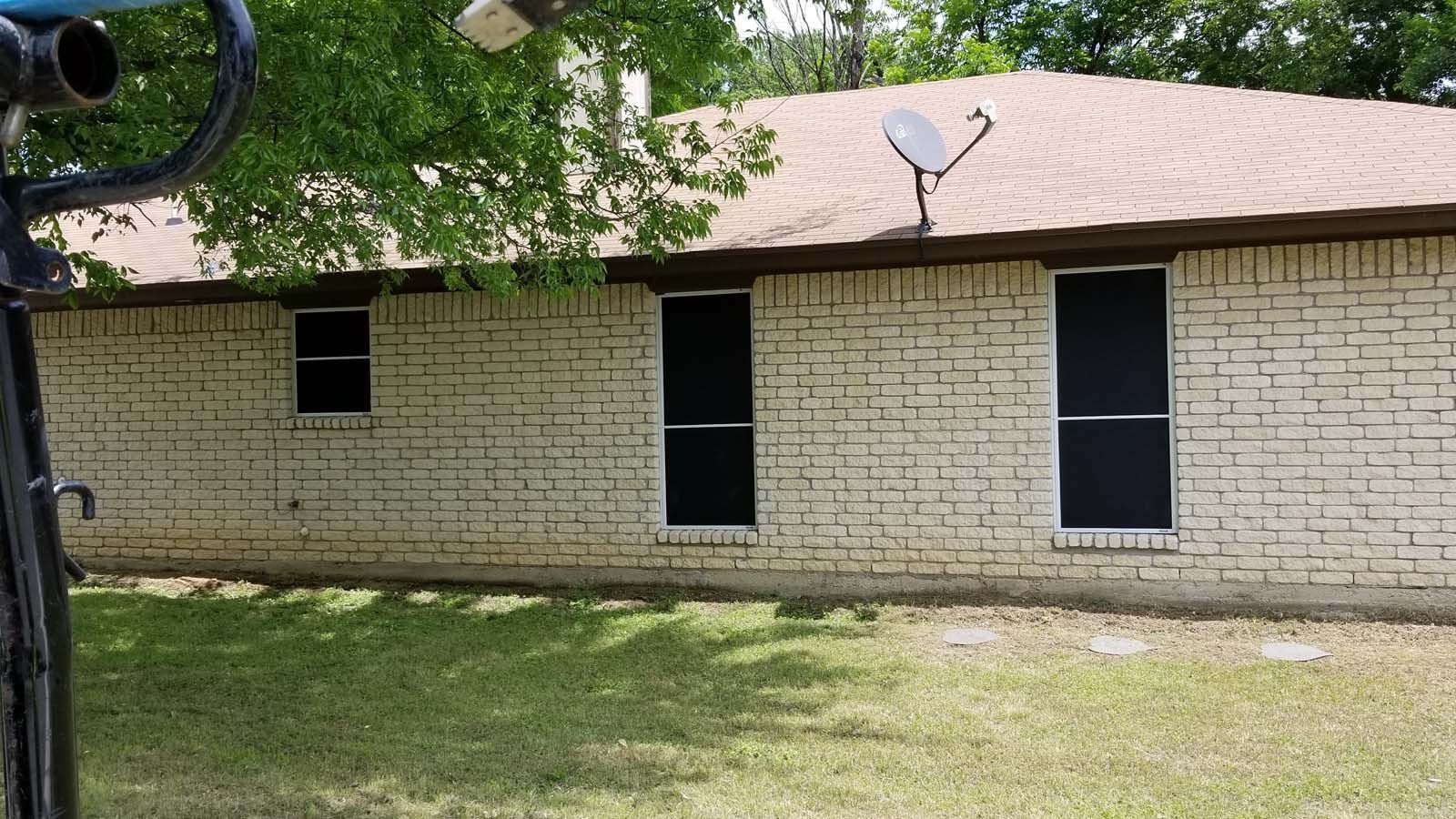 Tan brick house with dark window screens and satellite dish. Green grass and trees in the background.