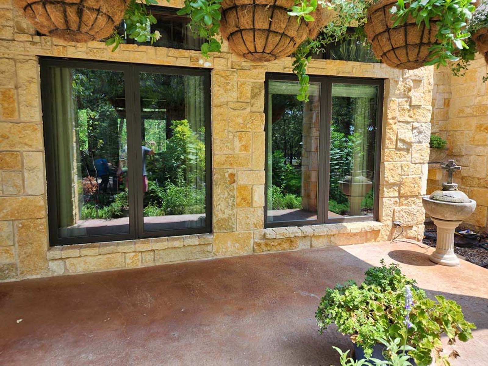 Two black-framed windows in a tan stone wall, reflecting a garden. Hanging baskets and a stone fountain are nearby.