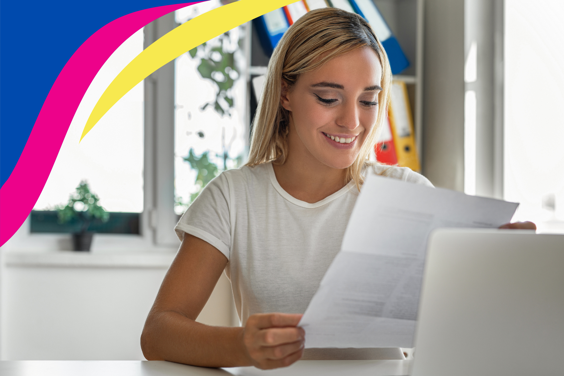 A woman is sitting at a desk looking at a piece of paper in front of a laptop computer.