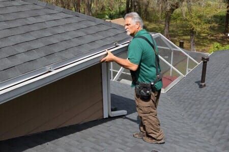 A man is standing on a roof looking at a gutter.