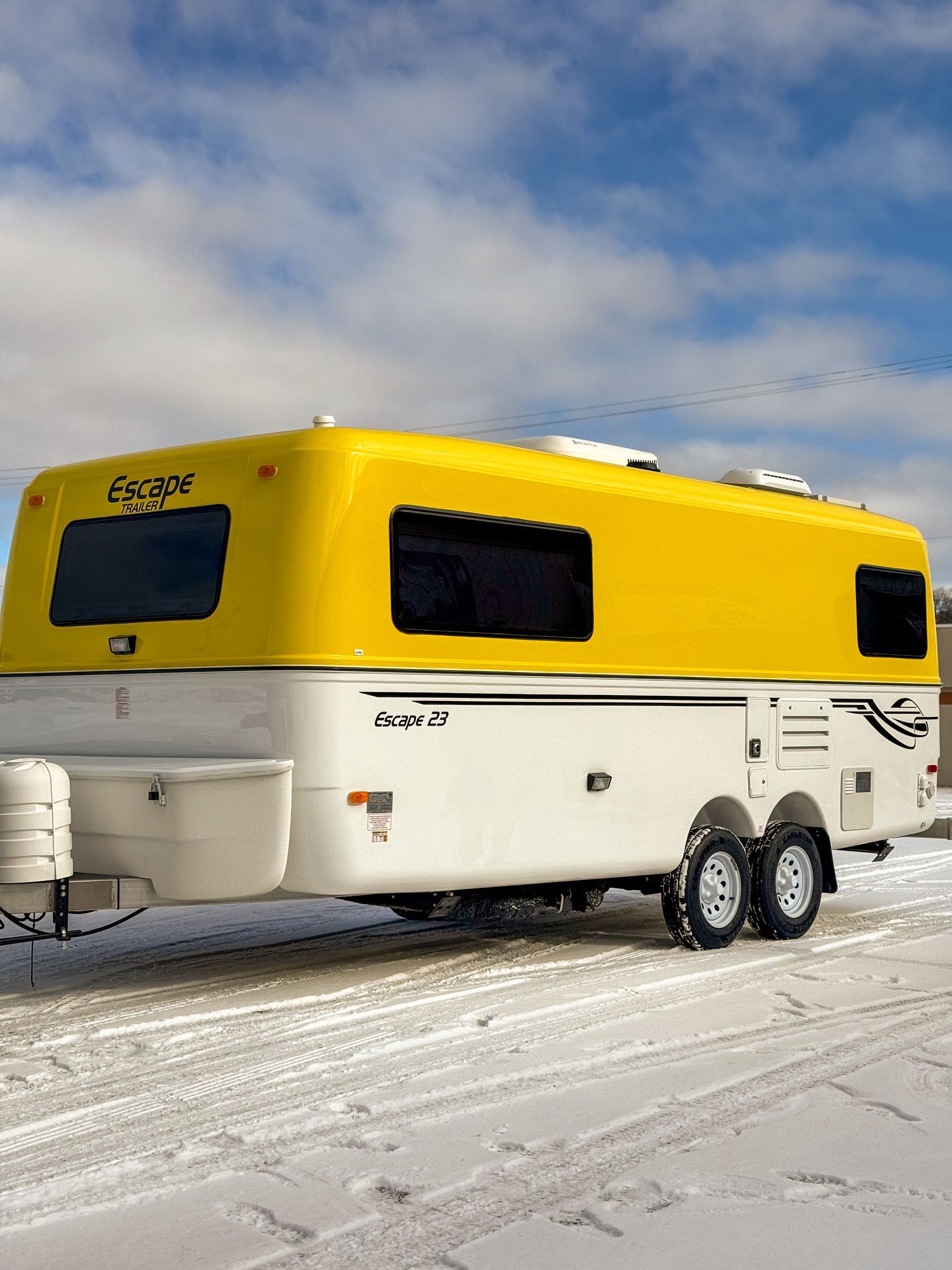 Yellow and white travel trailer with dual axles parked on snowy ground under a cloudy sky