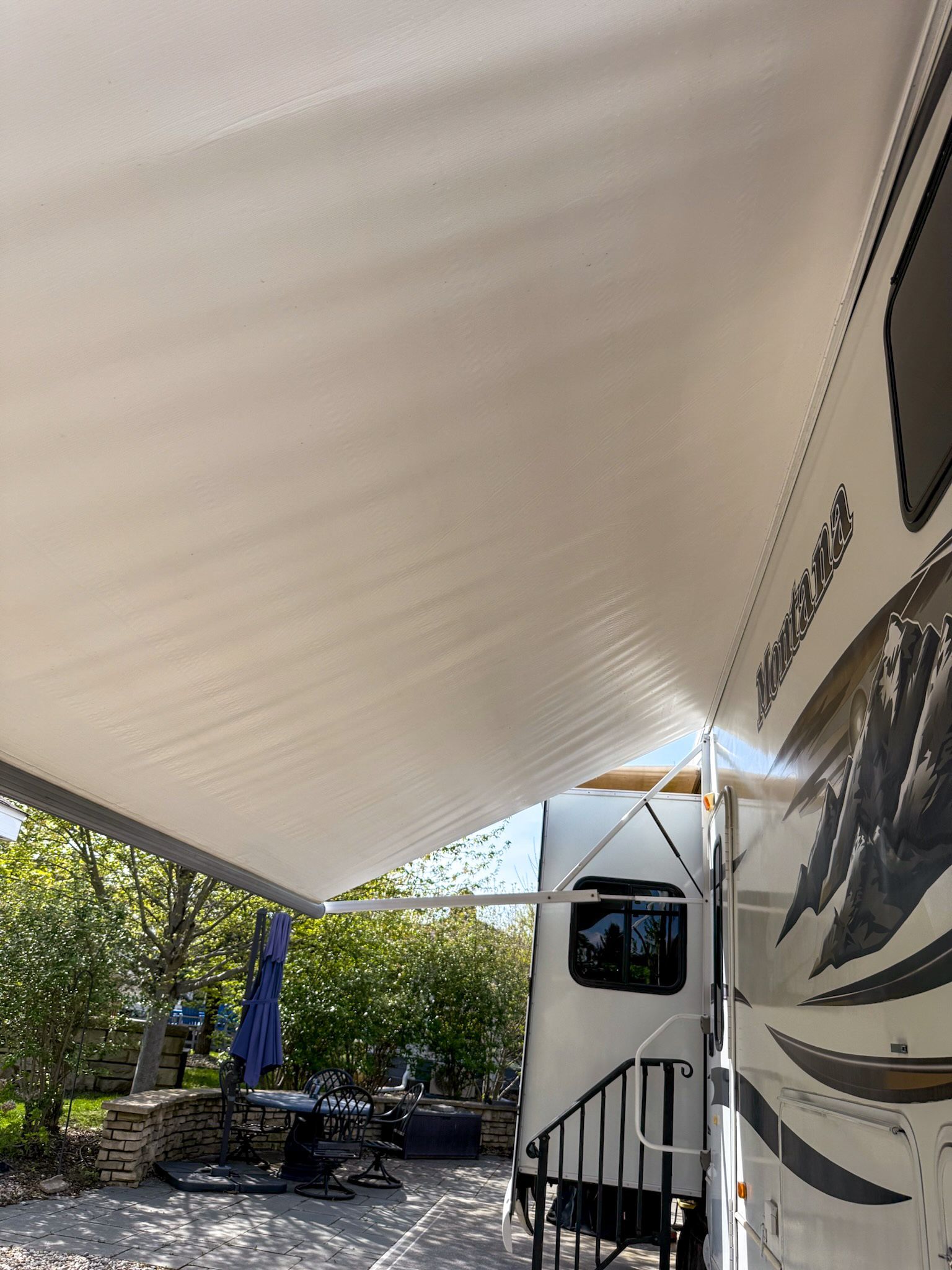 RV under a large white awning in a sunny campsite, with trees and outdoor furniture nearby