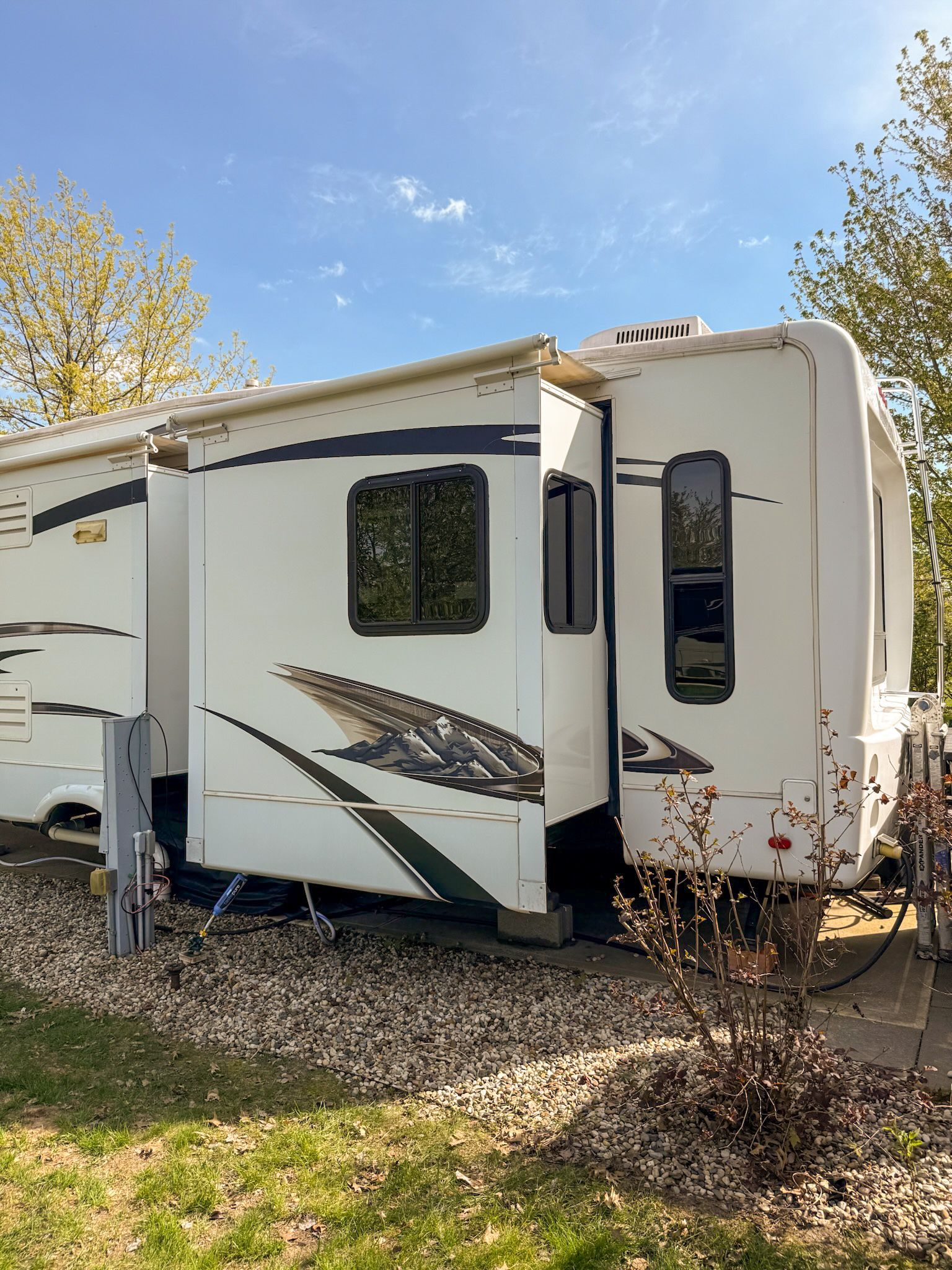 White travel trailer with a slide-out parked on grass under a blue sky, with autumn trees nearby