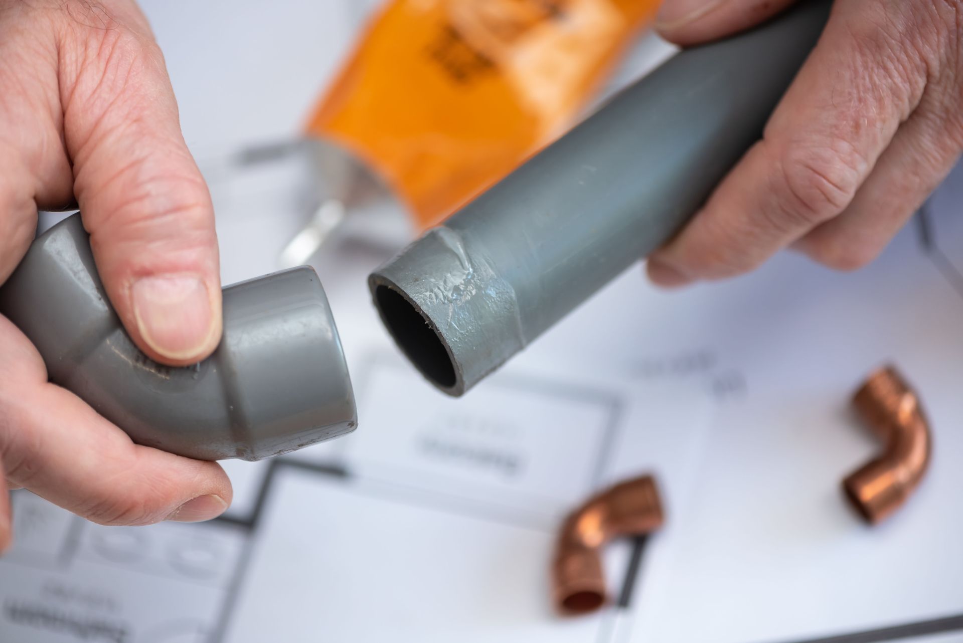 Hands fitting gray PVC pipe elbows on a workbench with copper fittings nearby