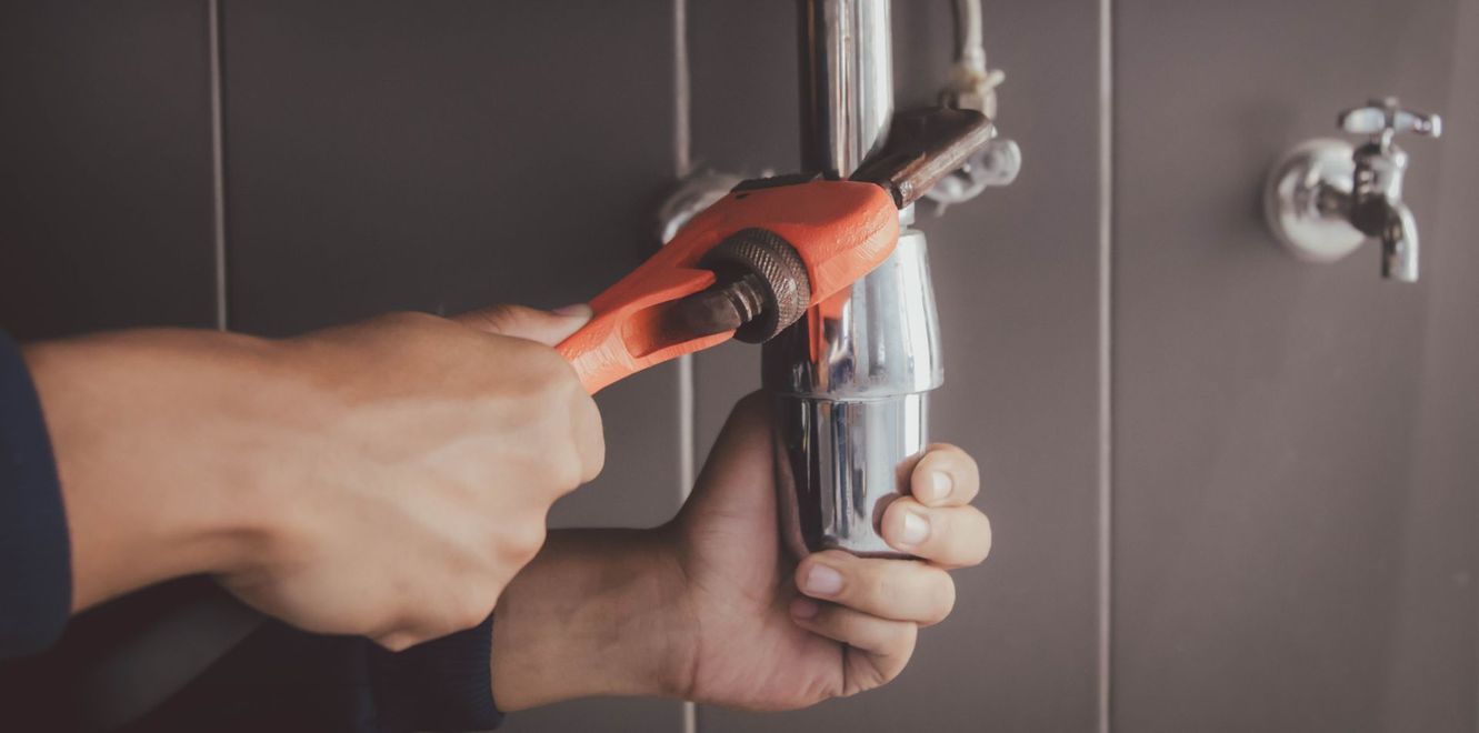 Hands tightening a chrome pipe with orange pliers under a sink cabinet