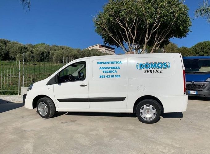 Un furgone di servizio bianco parcheggiato all'aperto accanto a un prato e a un albero alto, sotto un cielo azzurro e limpido.