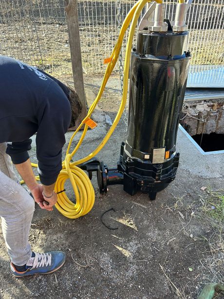 Un operaio avvolge un tubo giallo accanto a una pompa nera su un terreno ghiaioso vicino all'acqua.