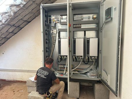 Un tecnico è inginocchiato accanto a un quadro elettrico aperto, intento a ispezionare il cablaggio sotto una scala.