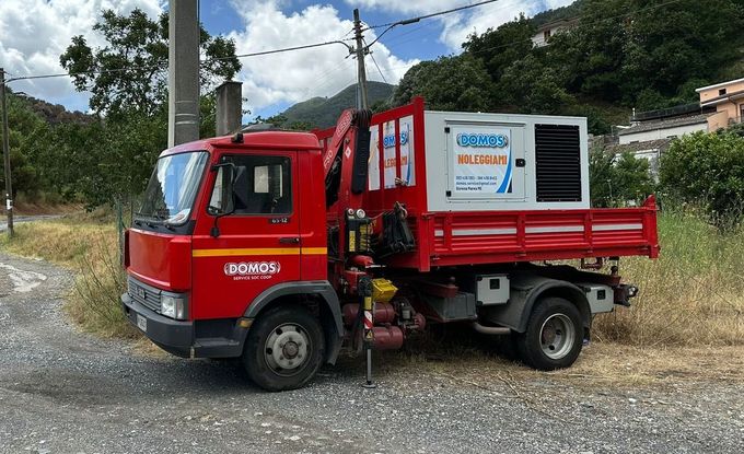 Camion rosso di servizio con attrezzature montate, parcheggiato sul ciglio di una strada sterrata vicino ad alberi e linee elettriche.