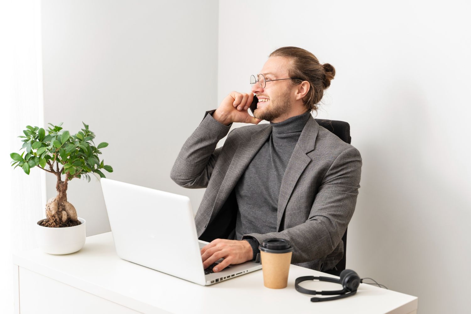 Person at a desk on a phone call, using a laptop with a coffee cup and potted plant nearby.
