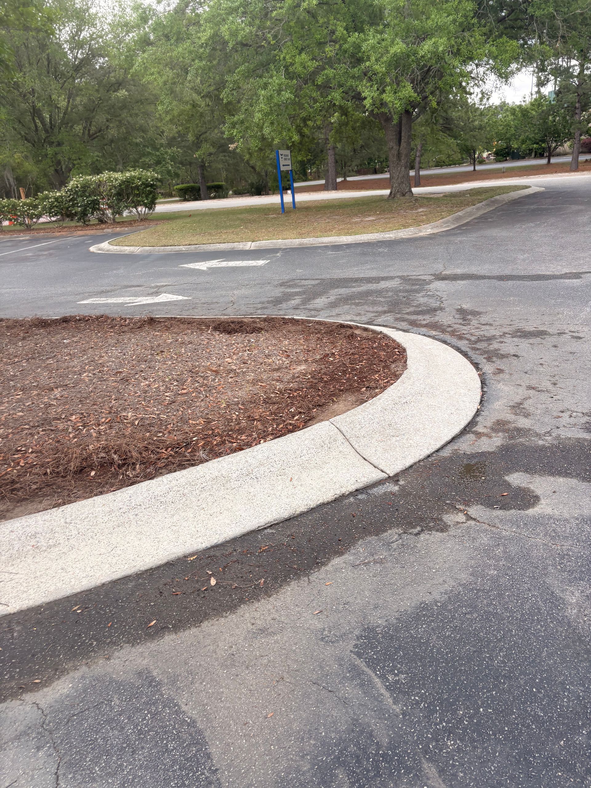 Curved curb and paved path in a wooded parking area with a blue sign in the background
