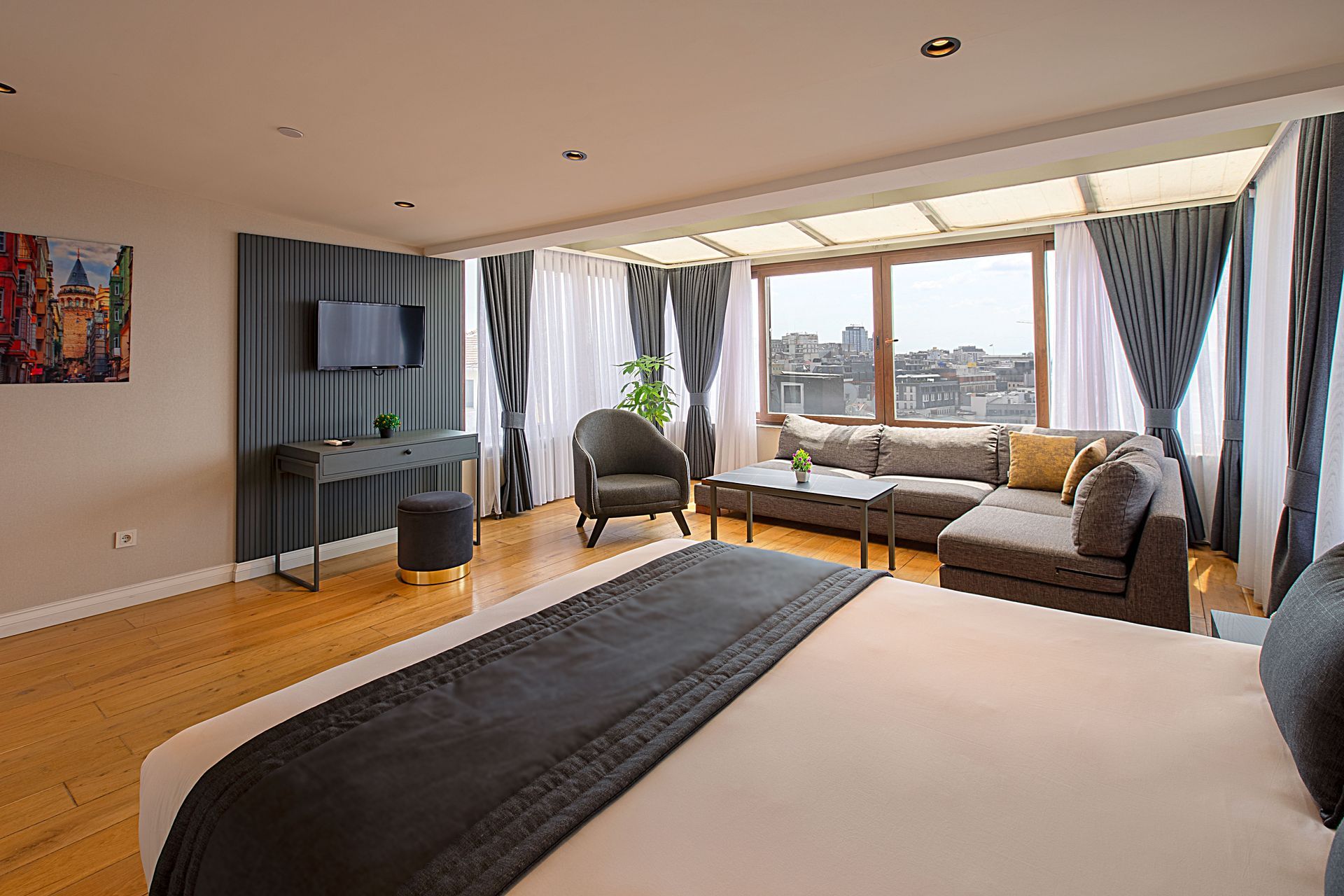 Yatak, oturma alanı, çalışma masası ve şehir manzaralı büyük pencerelere sahip geniş otel süiti.