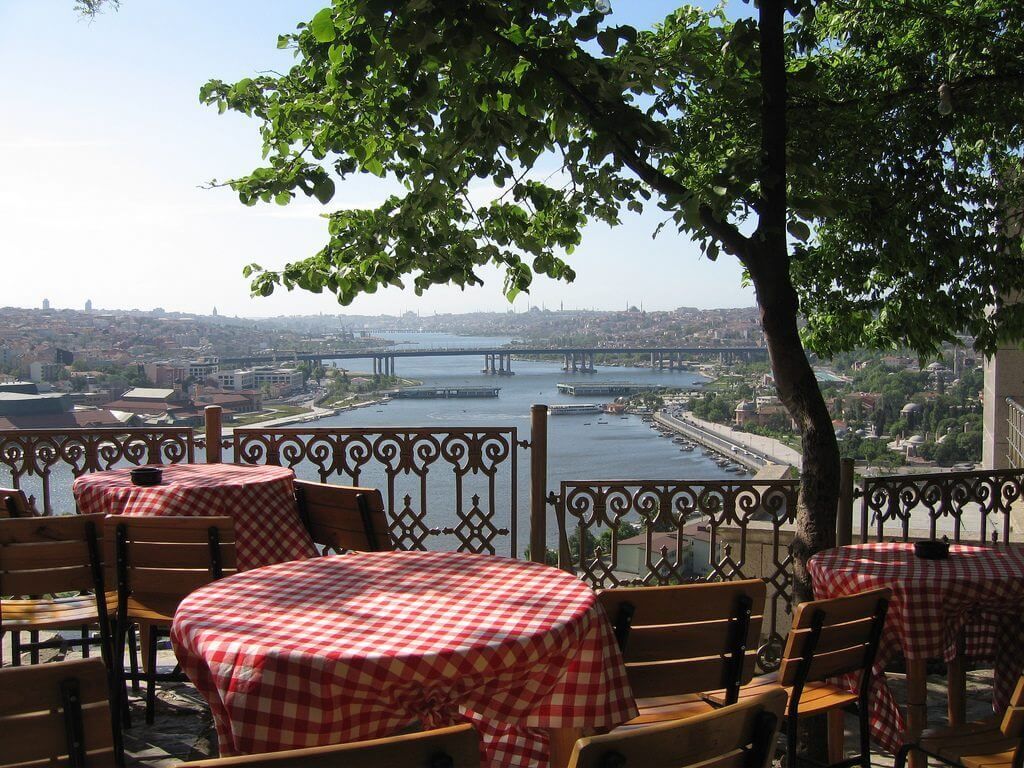 Kırmızı kareli masa örtülü restoran masaları, köprülü bir şehre ve nehre bakıyor. Manzarayı ağaç dalları çerçeveliyor.