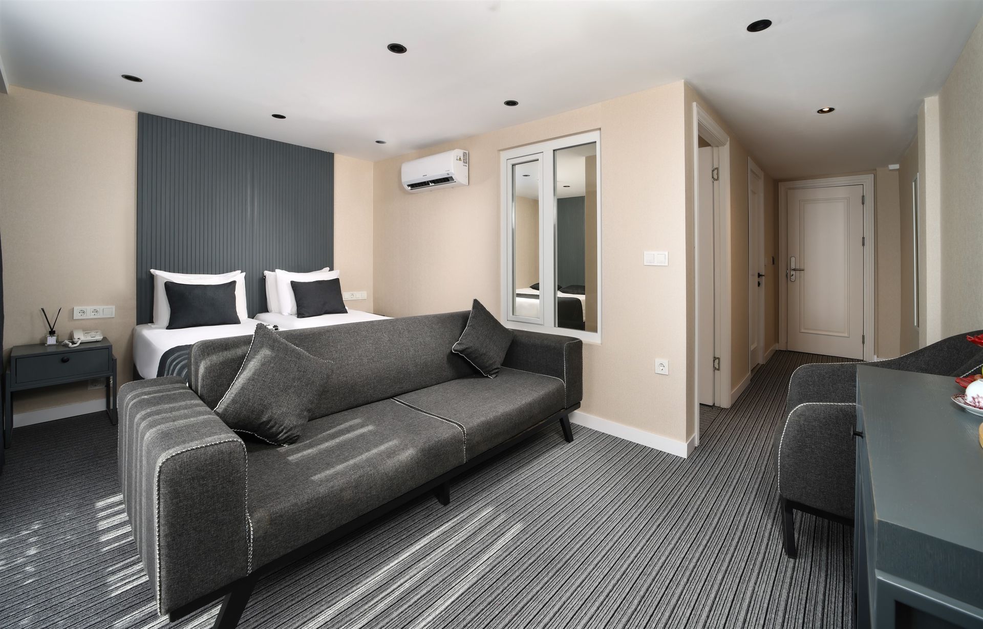 Gri kanepe, koltuk, yatak ve balkon kapıları bulunan otel odası. Ahşap zemin ve dekoratif aydınlatma.