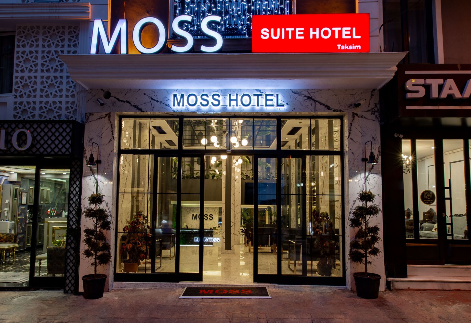 Entrance of MOSS Suite Hotel at night, with bright neon signage and glass doors illuminated inside
