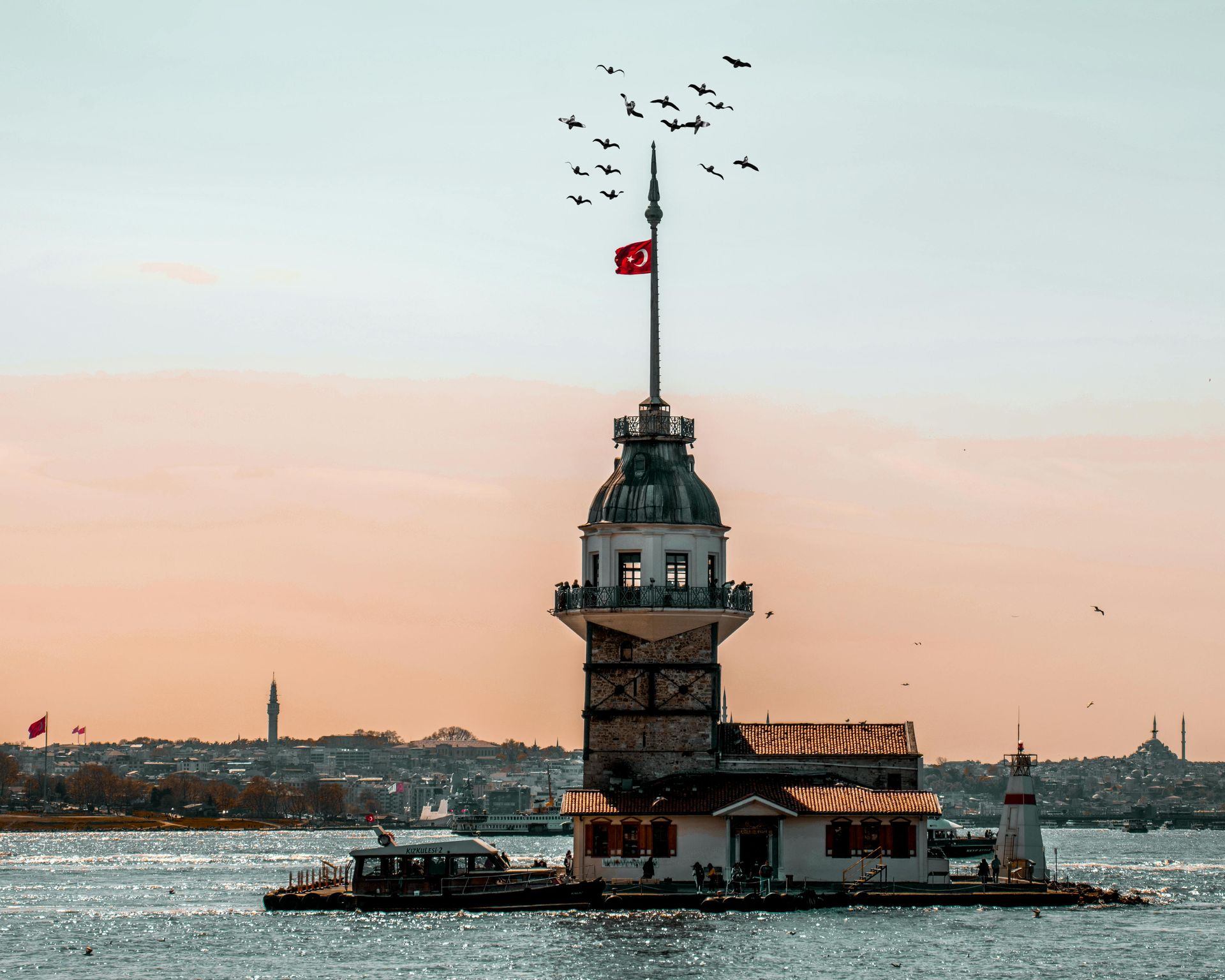 Türkiye, İstanbul'daki Kız Kulesi. Küçük bir adanın üzerinde beyaz kule, tepesinde Türk bayrağı, gökyüzünde uçan kuşlar.