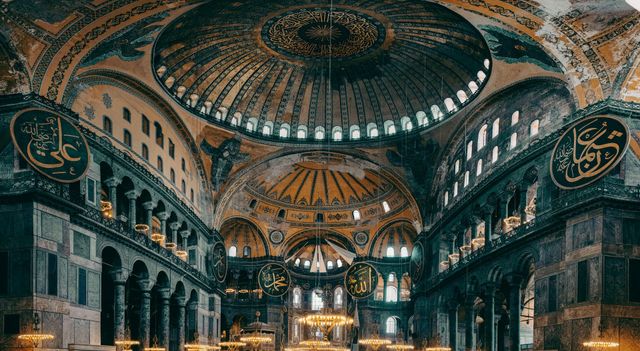 İstanbul Ayasofya'nın içi. Büyük kubbeli tavan, altın ve kahverengi detaylar, süslü mimari, avizeler ve sütunlar.