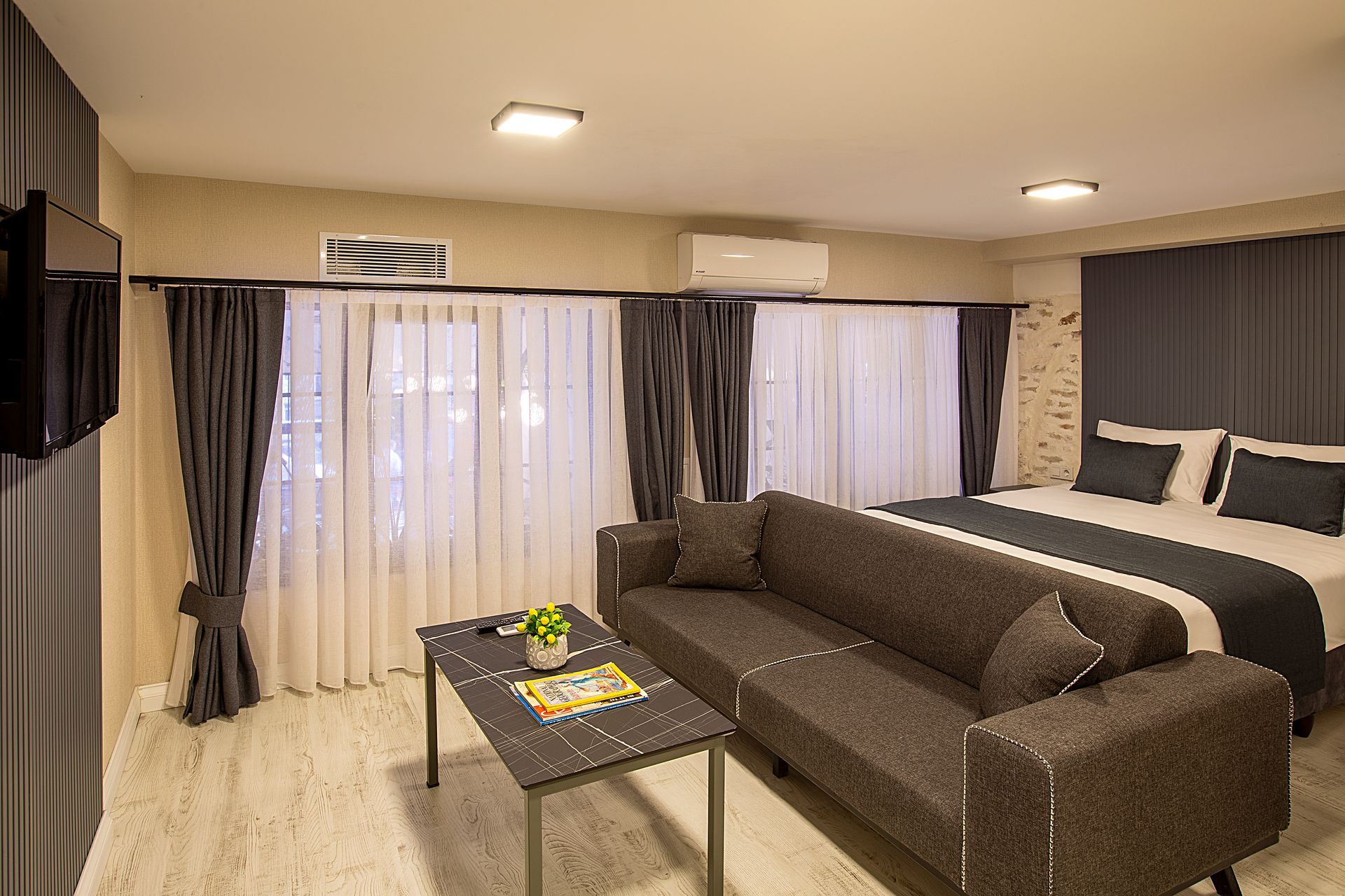 Modern living room with dark sofa, coffee table, curtains, and a bed in the background