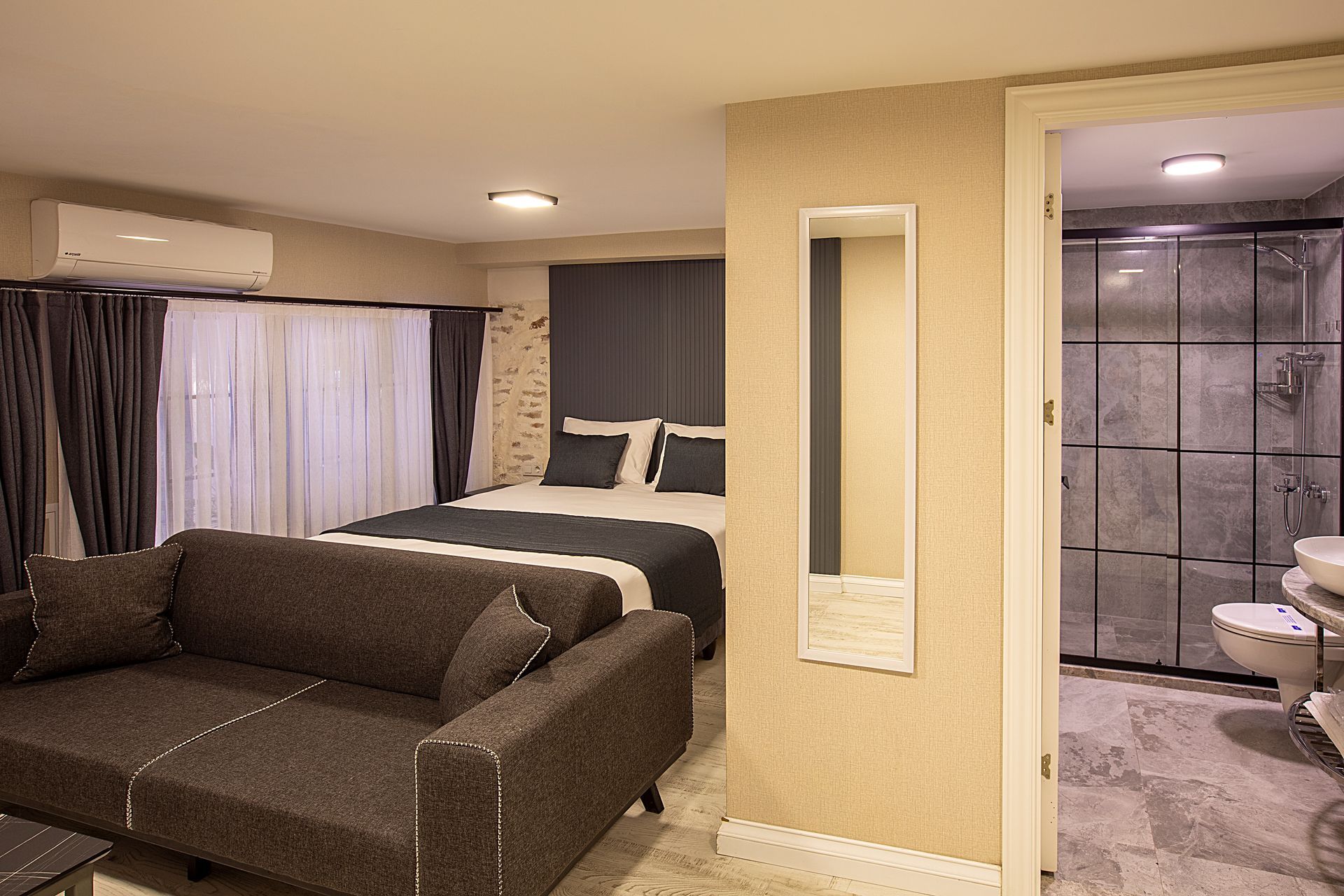 Modern bedroom suite with dark sofa, bed, curtains, and adjacent bathroom visible through an open doorway