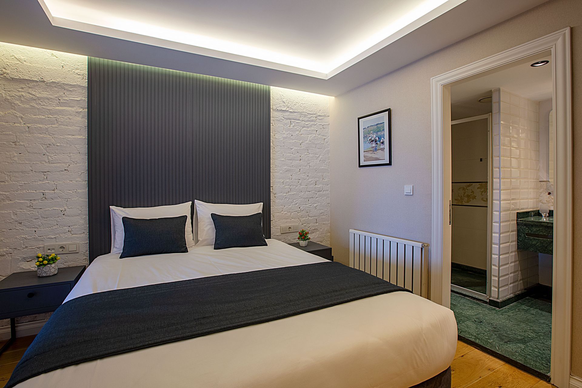 Modern bedroom with gray headboard, white brick walls, and an open doorway to a green-tiled bathroom